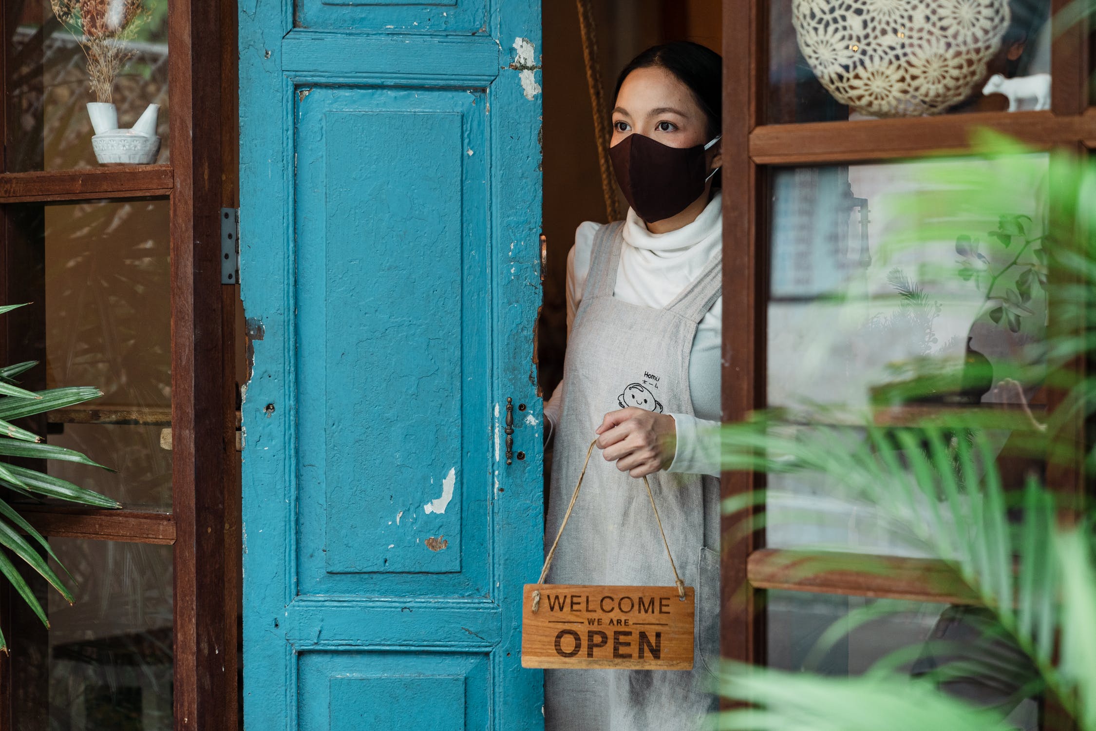 A woman wearing a face mask and apron stands in a doorway next to a blue door, holding a wooden “Welcome, we are open” sign. Green plants surround the entrance, creating a cozy and inviting atmosphere.