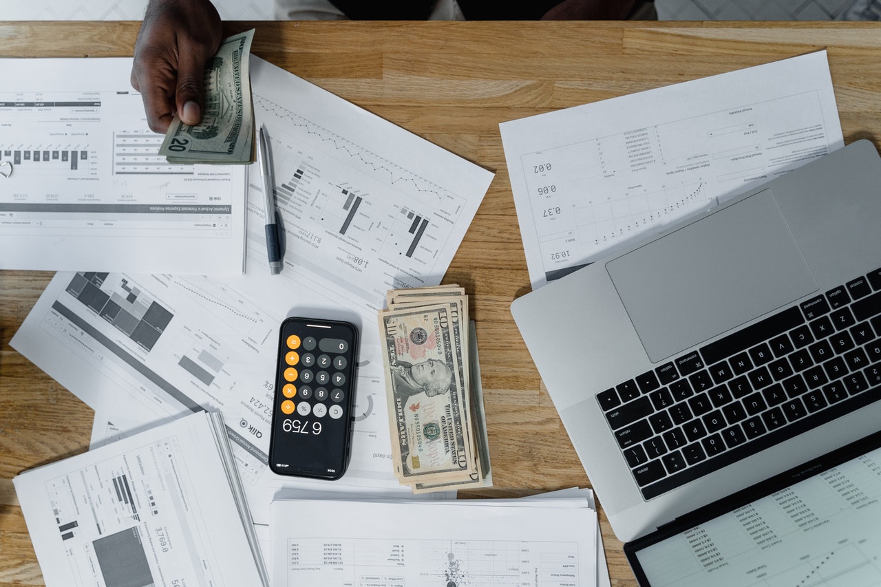 A person counts U.S. dollar bills at a desk cluttered with financial documents, a pen, a laptop, and a smartphone displaying a calculator app. More bills are stacked beside the phone.