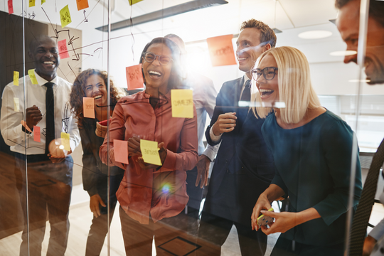 A group of colleagues smiling and brainstorming together in an office, standing around a glass wall covered with colorful sticky notes and sketches, sunlight streaming in behind them.