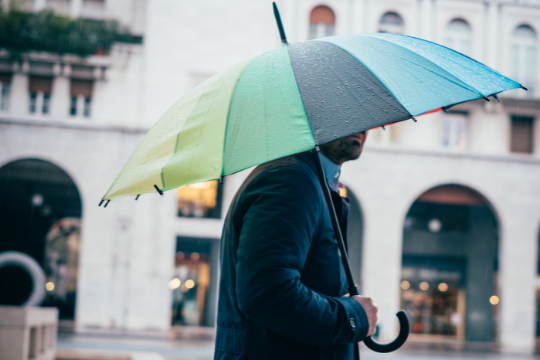 A person in a dark coat holds a colorful umbrella while walking on a rainy city street with arched buildings in the background.