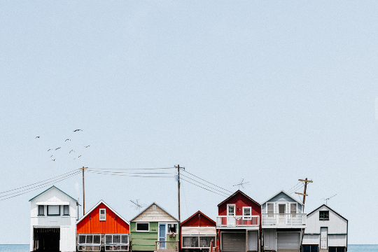 A row of colorful beach houses on stilts stands along the shoreline under a clear blue sky, with several birds flying above and power lines stretching across the scene.
