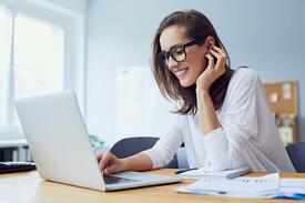 A woman wearing glasses and a white top is smiling while working on a laptop at a desk. She has a notepad and documents nearby and appears to be in a bright, modern office.