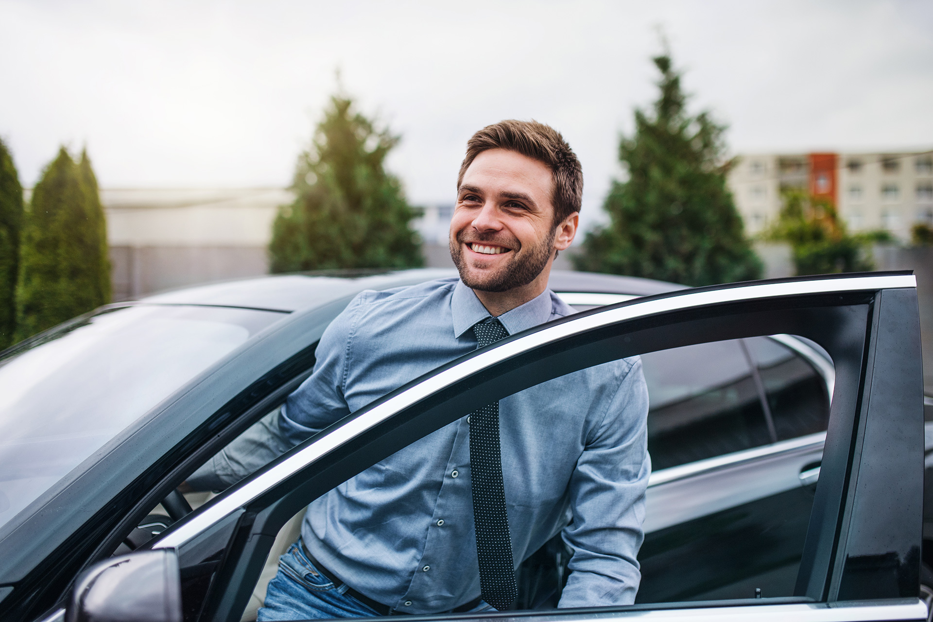A smiling man in a blue shirt and tie is stepping out of a car with the driver’s door open, perhaps heading to discuss remote work tax deductions. Trees and buildings are visible in the background on this sunny day outdoors.