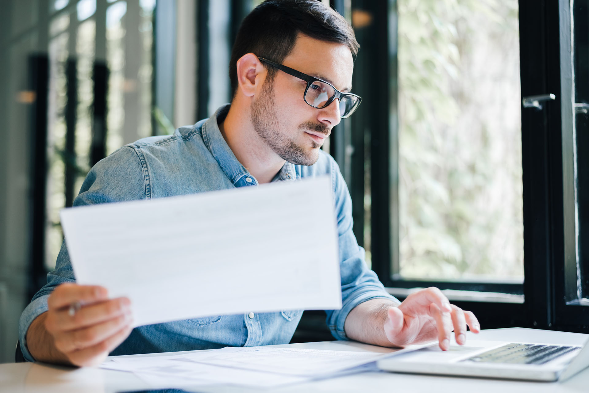 A man wearing glasses and a denim shirt sits at a desk, looking at a laptop while holding a sheet of paper. He appears focused, with large windows and greenery visible in the background.