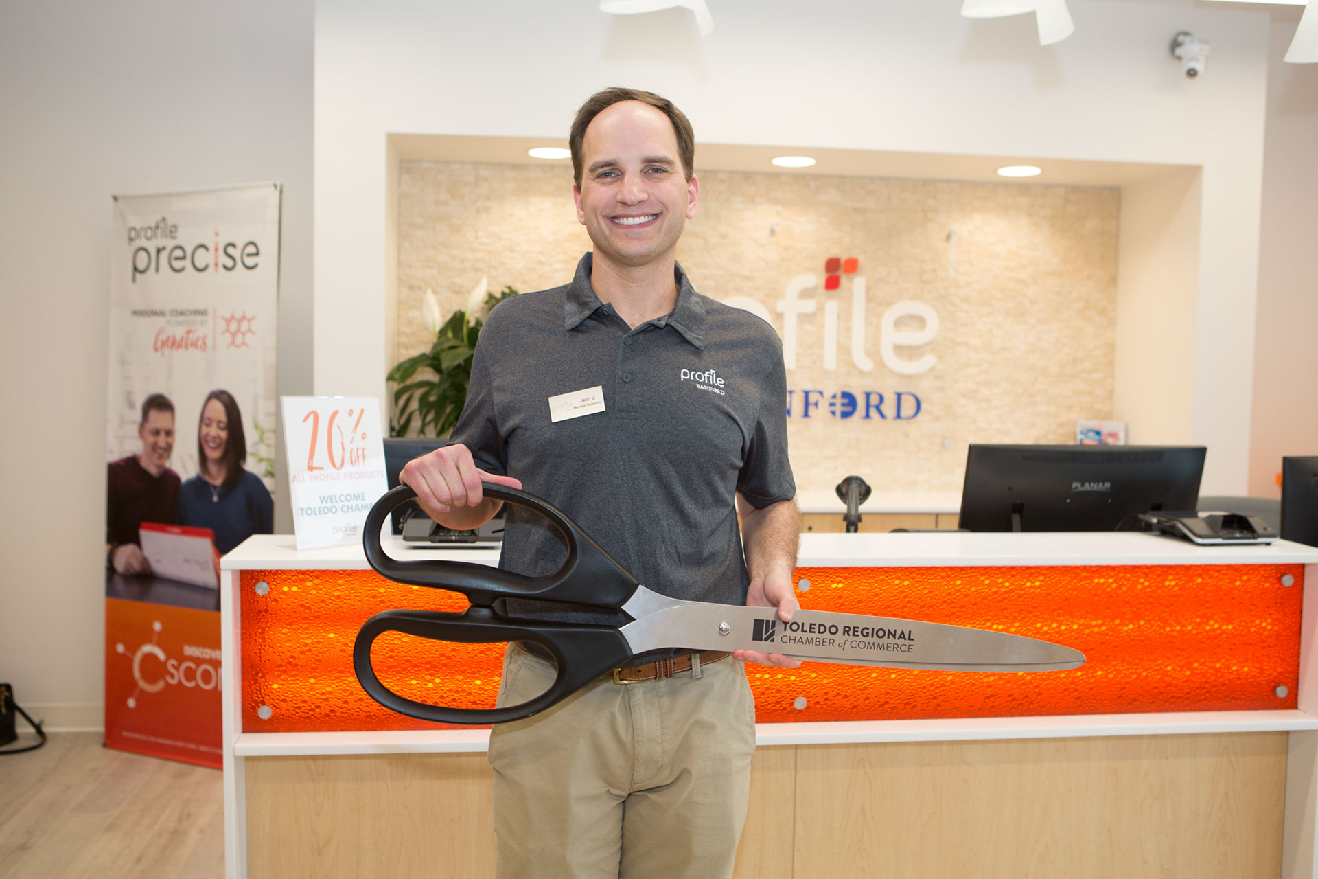 A smiling business owner stands in an office lobby holding a giant ceremonial pair of scissors. He wears a name tag and polo shirt, with a reception desk and promotional sign visible in the background.