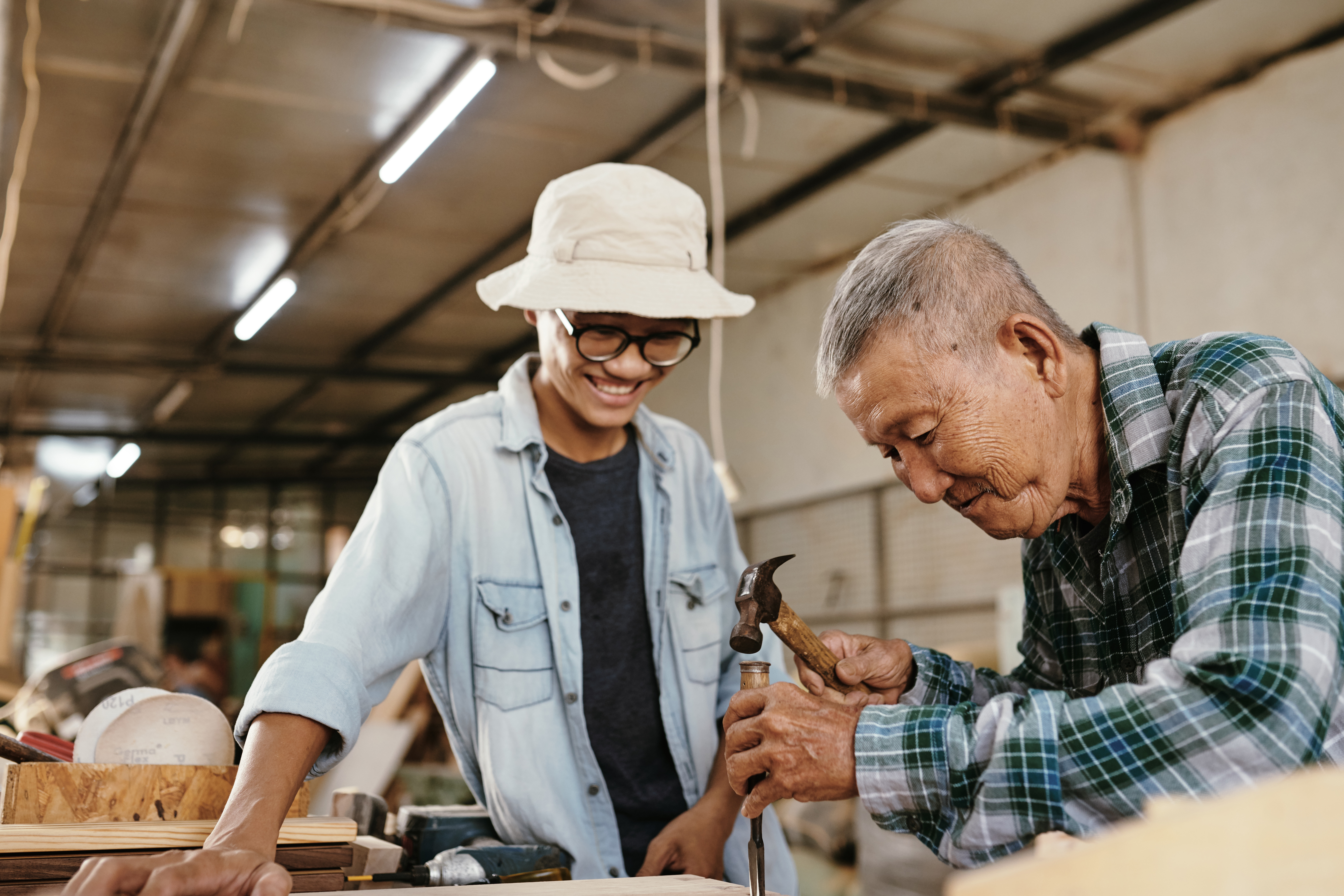 An older man uses a hammer on a piece of wood while a younger man in glasses and a hat smiles and watches, capturing the joy of working with family in a workshop filled with tools and materials.