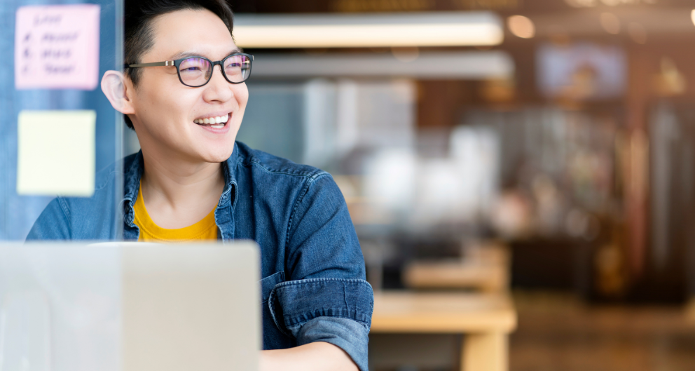 A young Asian man sits at his laptop, working as a remote employee. He smiles at something off in the distance.