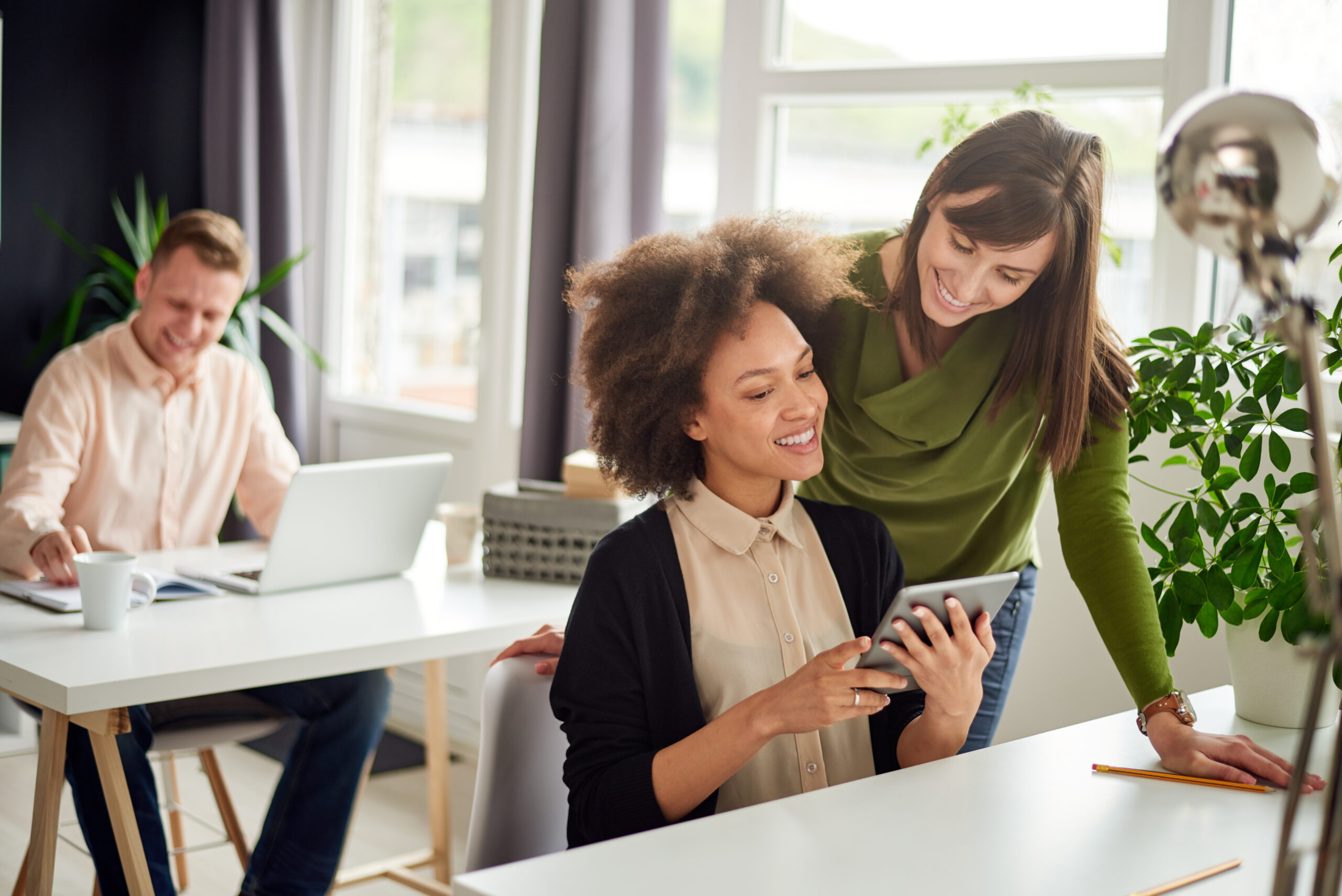 Two women smiling and looking at a tablet together in a bright office, possibly discussing payroll partner solutions, while a man works on a laptop in the background. The office features large windows, plants, and white desks.