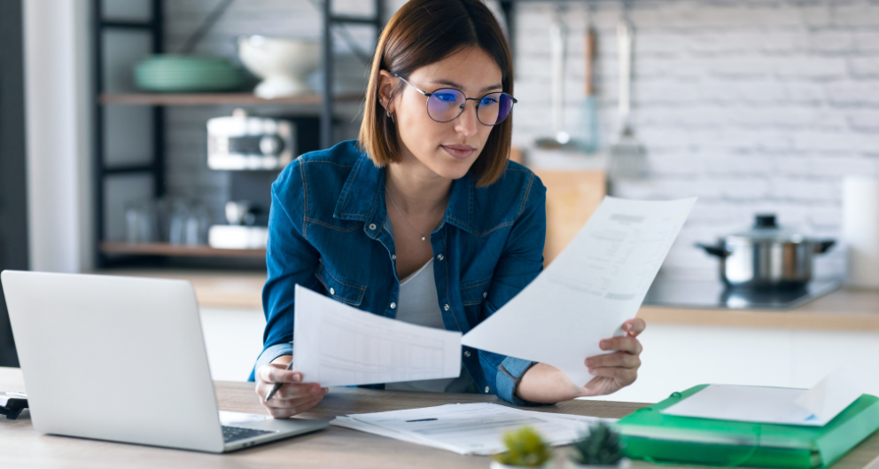 A young woman sits at her kitchen counter with a laptop and a stack of papers, working on business tasks.
