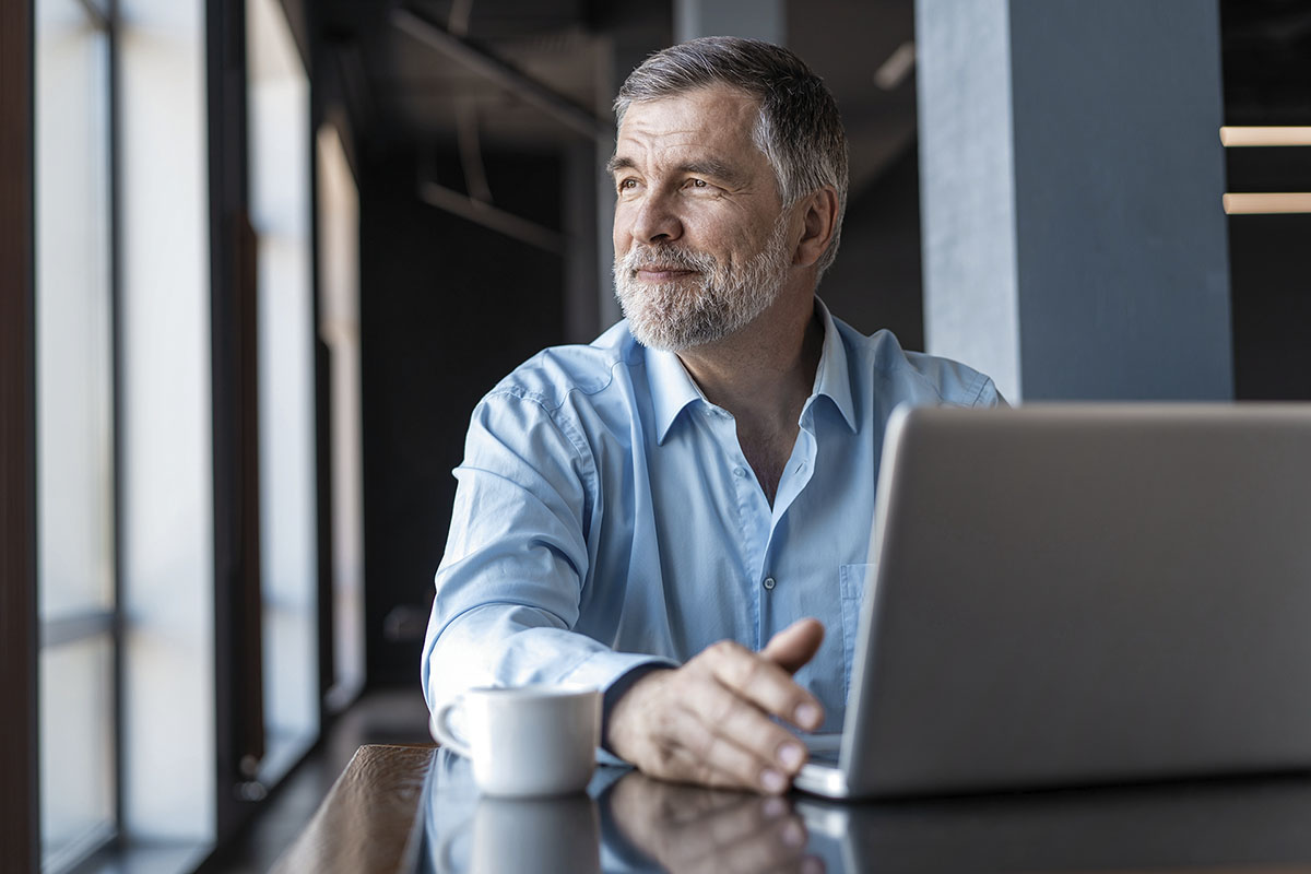 Older man with gray hair and beard, wearing a light blue shirt, sits at a table with a laptop and coffee cup, looking thoughtfully out of a window in a bright, modern setting.