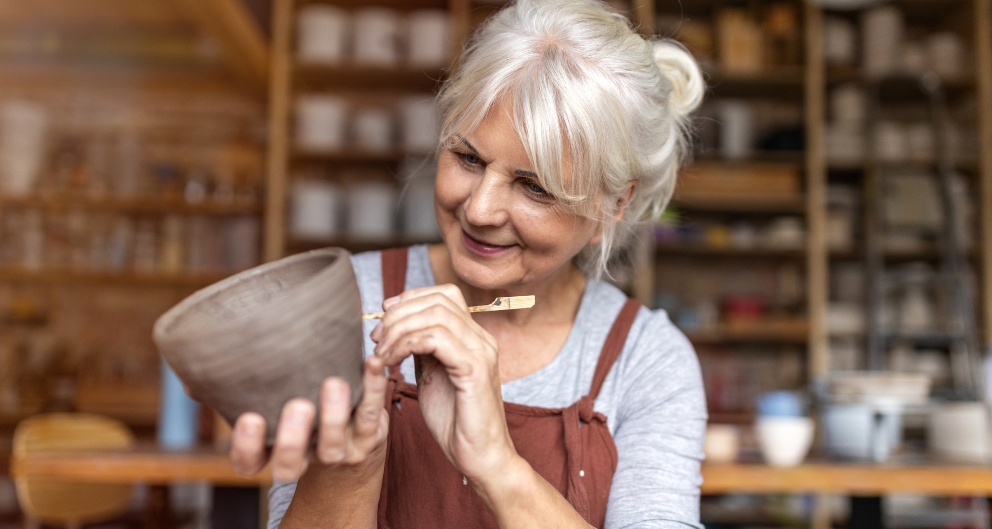 An older woman wearing an apron sits in a pottery studio, holding up a clay bowl as she carves designs into it