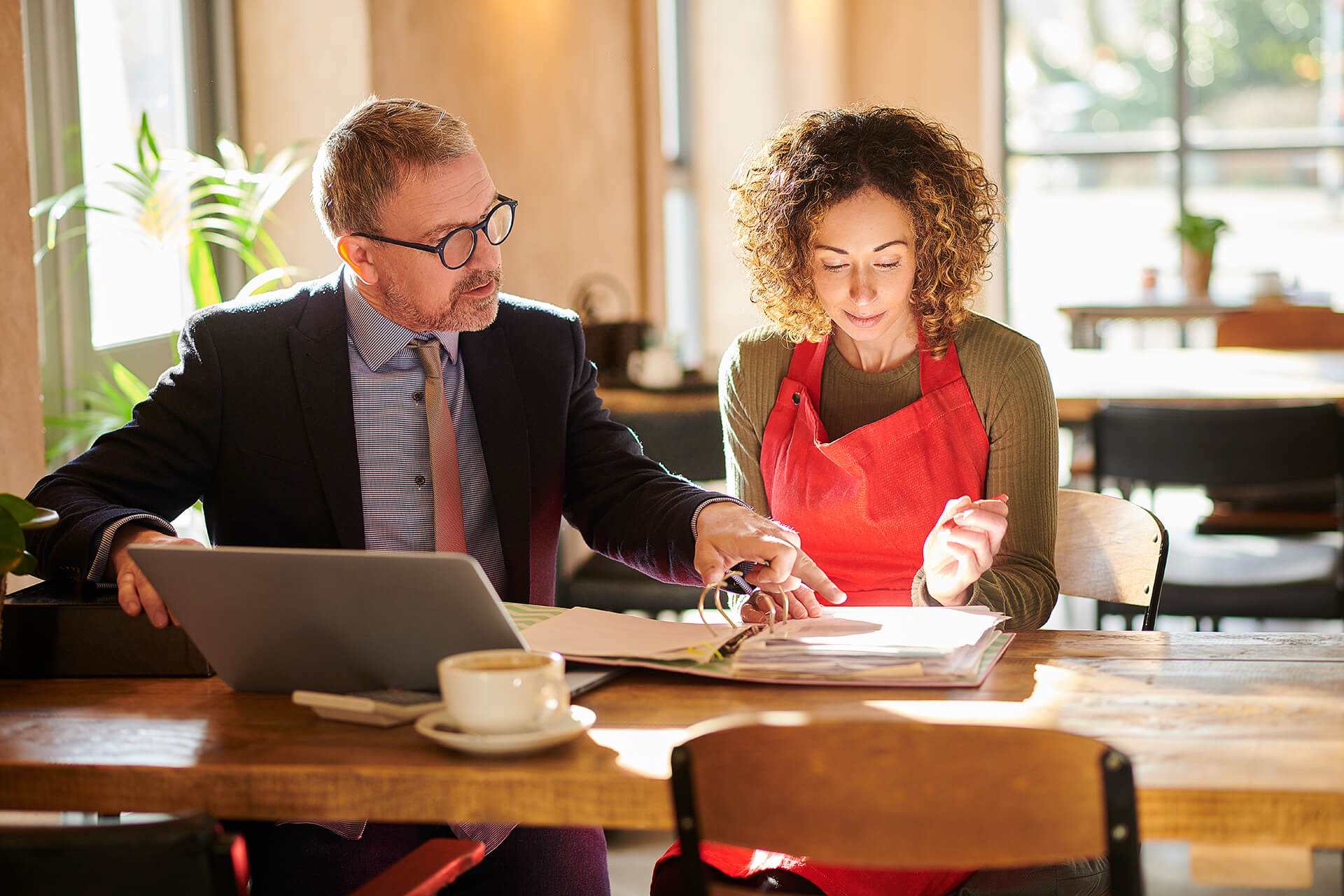 A man in a suit and a woman in a red apron sit at a table in a bright cafe, reviewing documents together; a laptop and coffee cup are on the table.