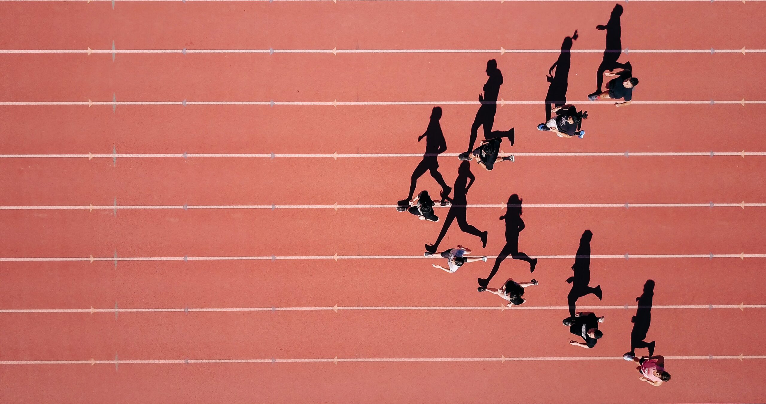 Aerial view of several runners on a red Padgett track, casting long shadows as they run in different lanes. The image captures the motion and spacing of the athletes from above.
