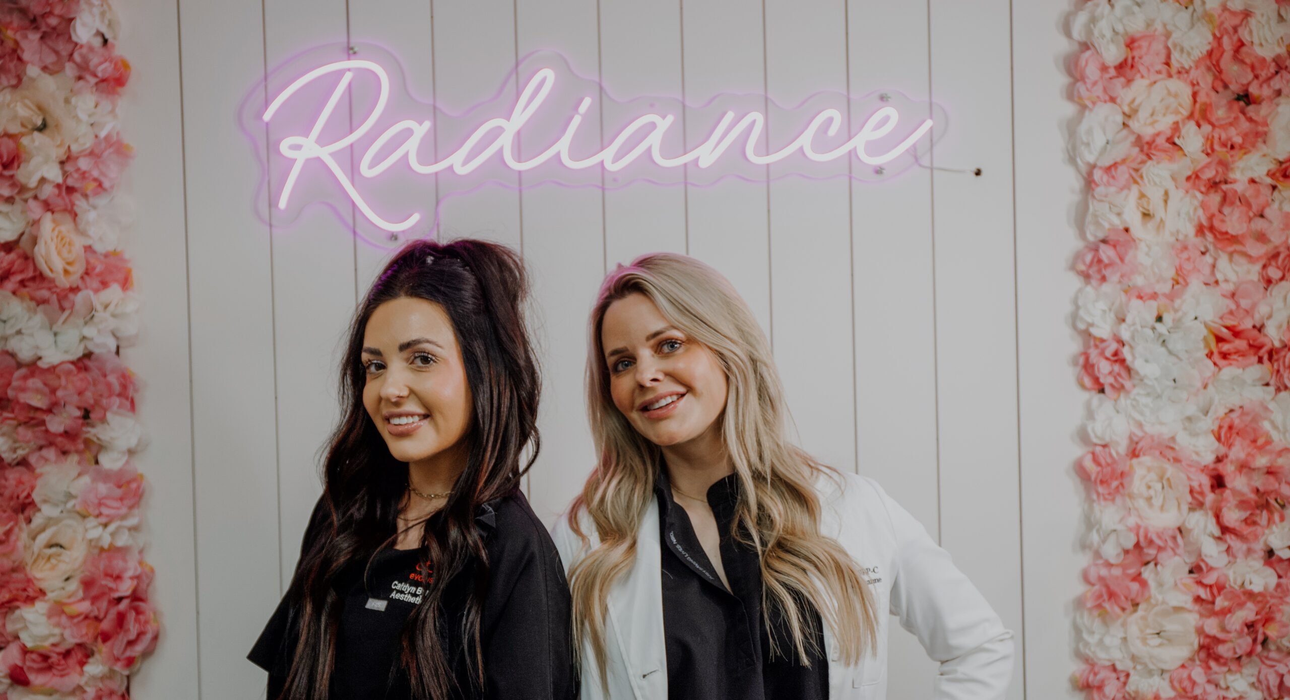 Two women smiling and standing in front of a pink neon Padgett Radiance sign, with floral decorations on the white paneled wall behind them.