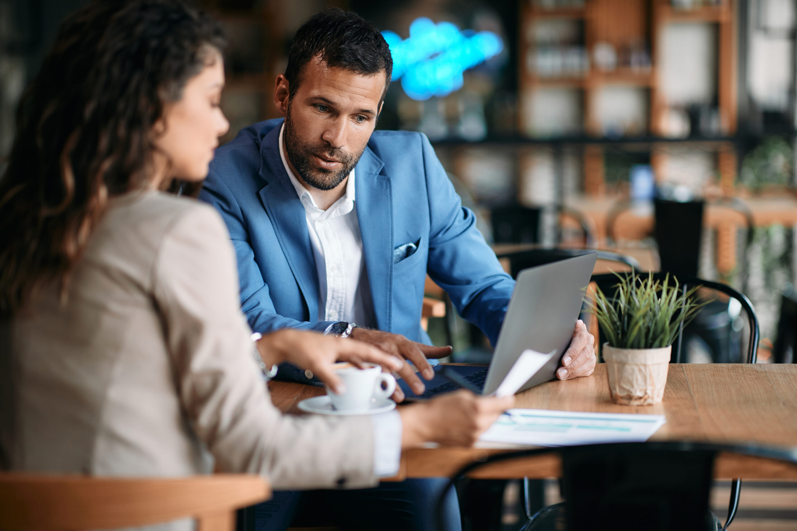 A man in a blue suit and a woman in a beige jacket discuss social security documents at a table in a cafe, with a laptop, coffee cup, and small plant in front of them.