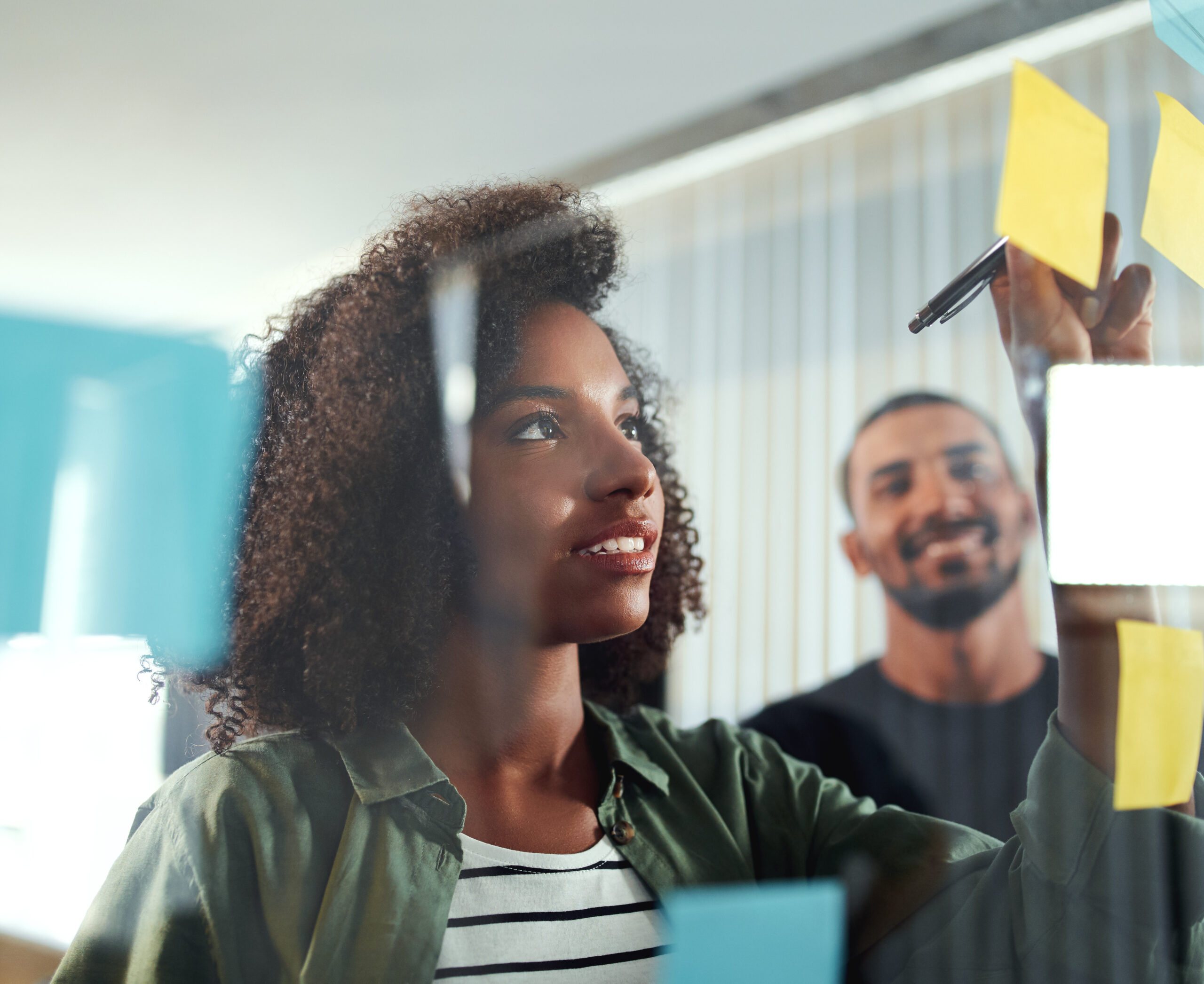 A woman writes on a yellow sticky note about cost segregation on a glass wall, holding a pen, while a smiling man stands in the background. Other sticky notes are also visible on the glass.
