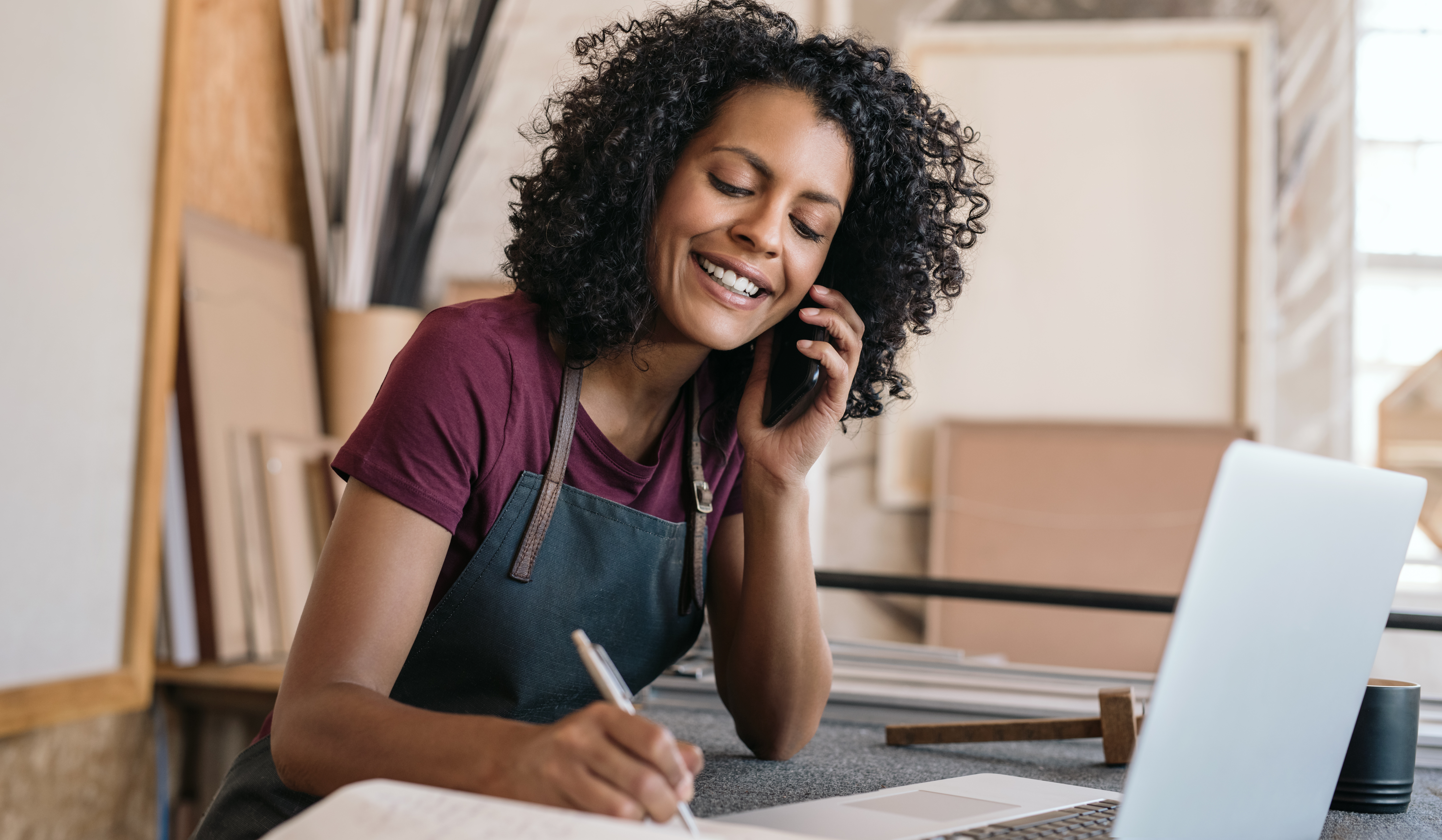 A woman with curly hair, wearing an apron, smiles while talking on the phone about 401(k) options and writing in a notebook at a desk with a laptop in a bright, modern workspace.