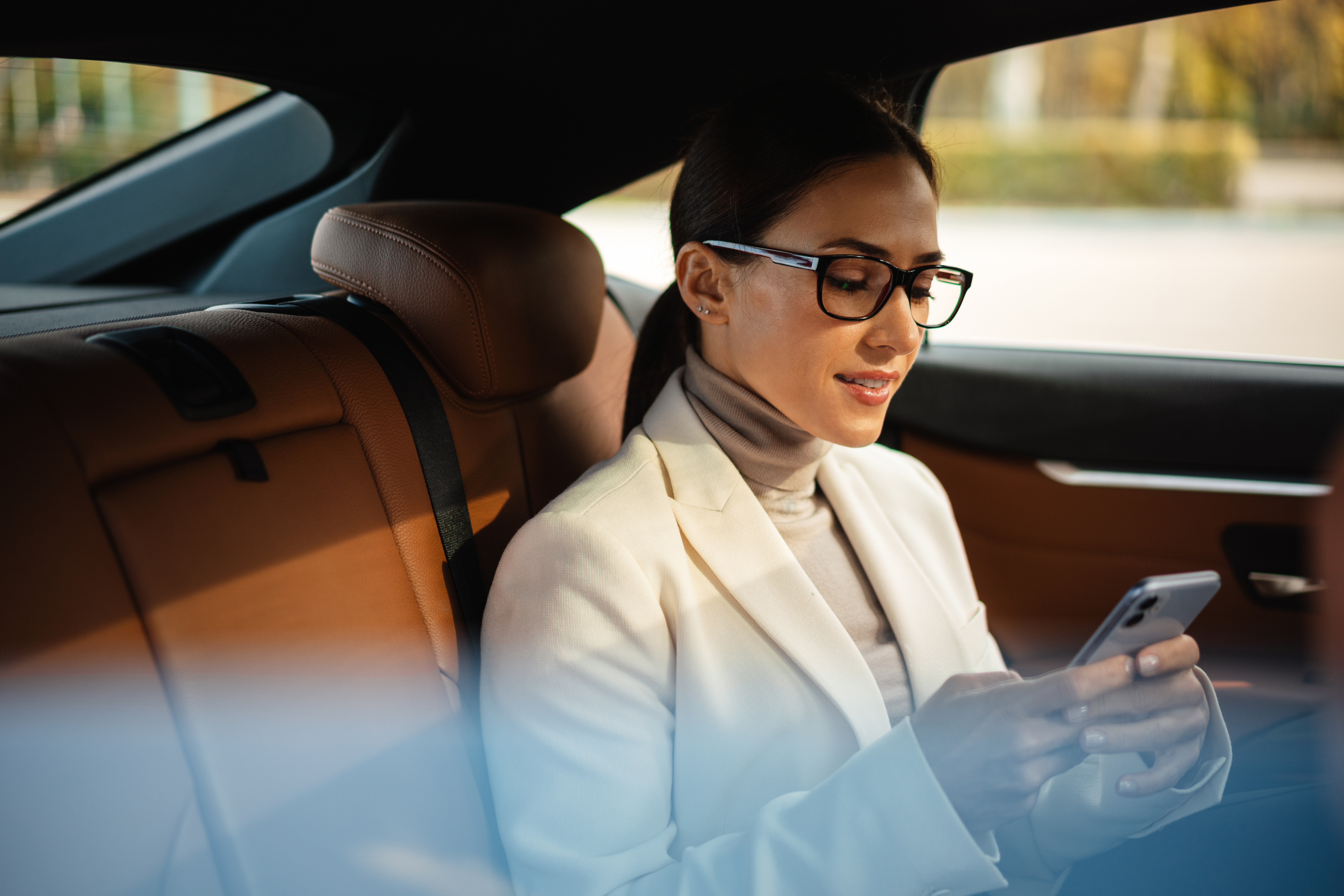 A woman wearing glasses and a white blazer sits in the back seat of a car, looking at her smartphone. She appears focused, possibly reviewing per diem expenses, and is surrounded by brown leather seats.