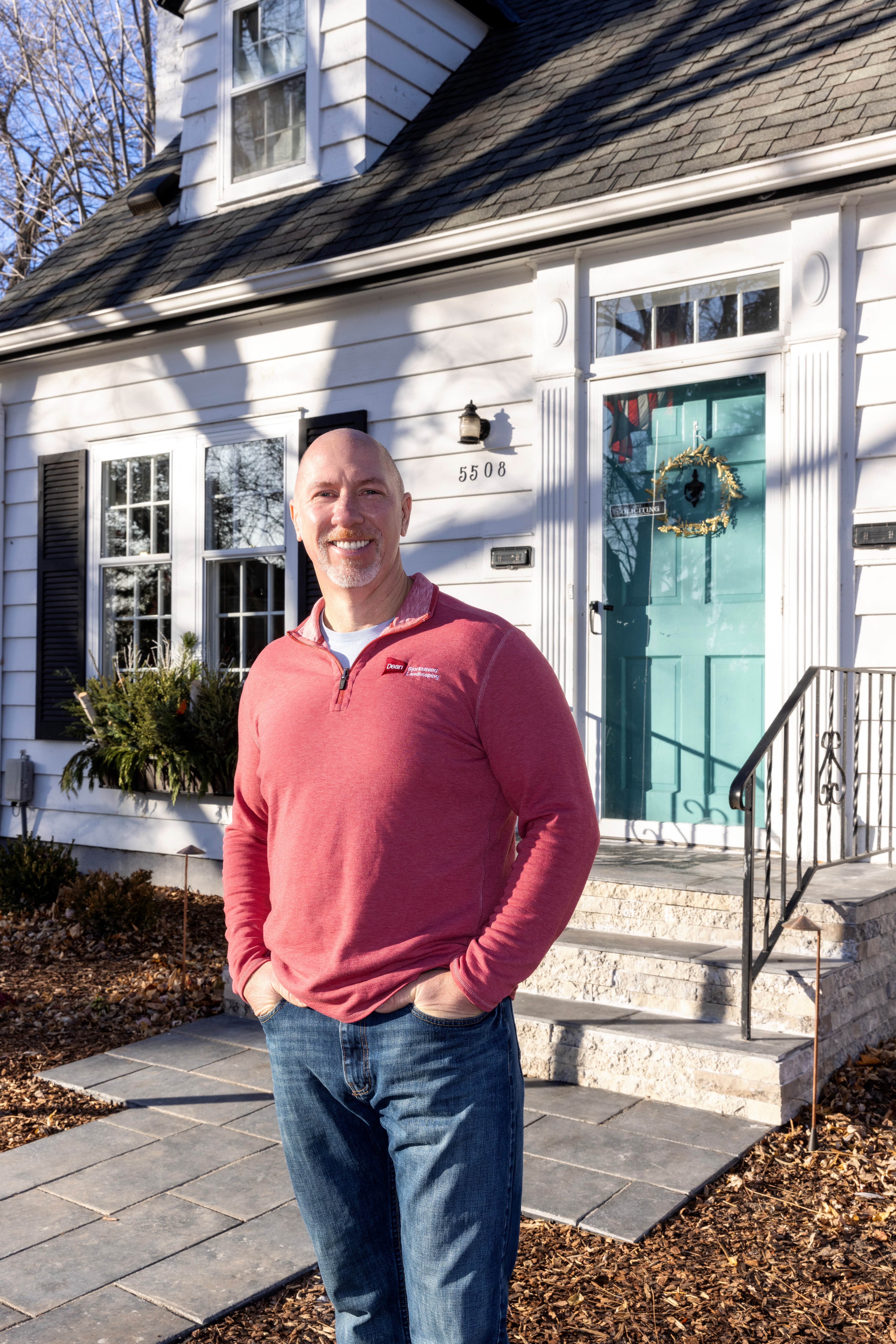 A man in a red pullover and jeans stands smiling in front of the Padgett house, a white home with black shutters and a turquoise front door adorned with a wreath. It is sunny, and the house number 5506 is visible.