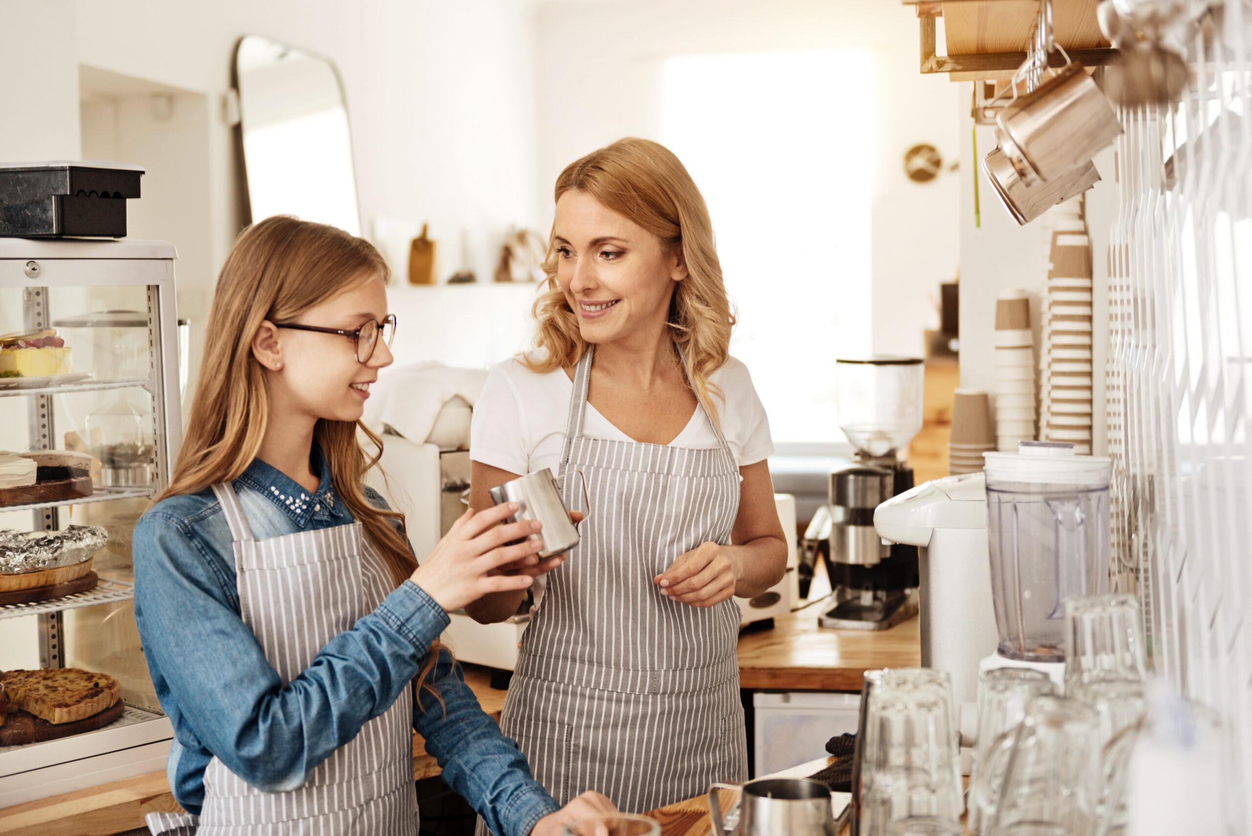 Two women wearing striped aprons stand behind a coffee shop counter. One, with glasses, holds a metal milk frothing pitcher, while the other smiles at her. Coffee equipment and kiddie treats are visible in the background.