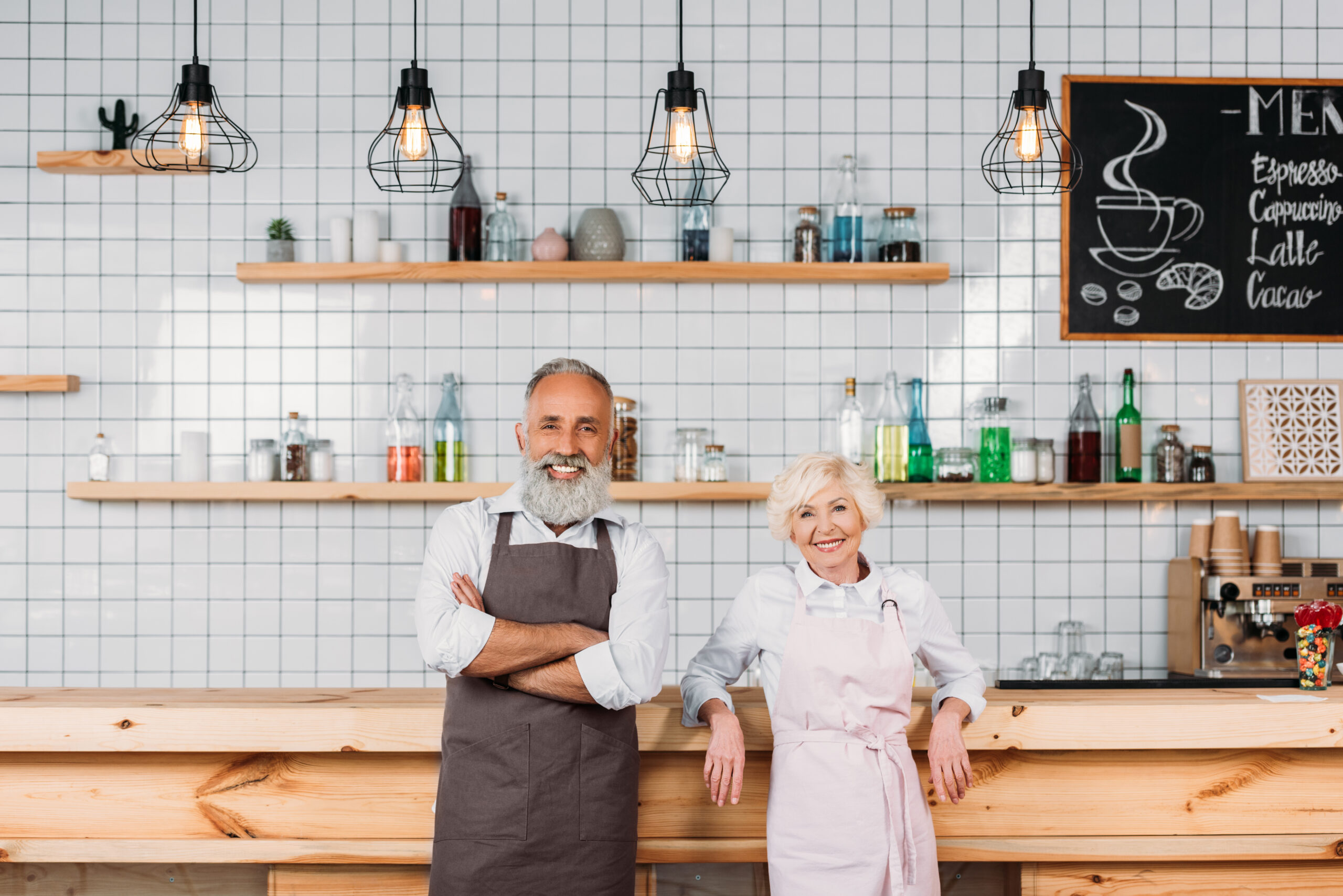 Two smiling older adults wearing aprons stand in a modern café with wooden counters, white tiled walls, and hanging lights above—ready to share friendly service and helpful tips.