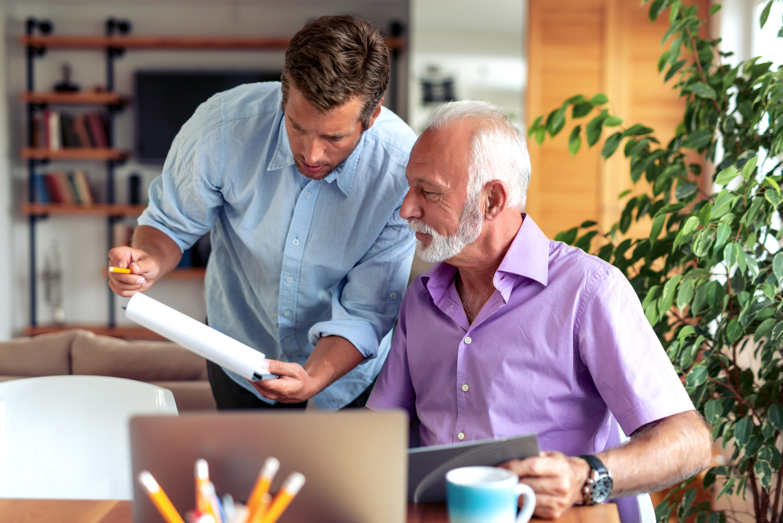 A younger man stands next to an older man seated at a table, both looking at documents. The table has a laptop and pens, creating a standard office feel, with shelves and plants in the background suggesting a casual workspace.