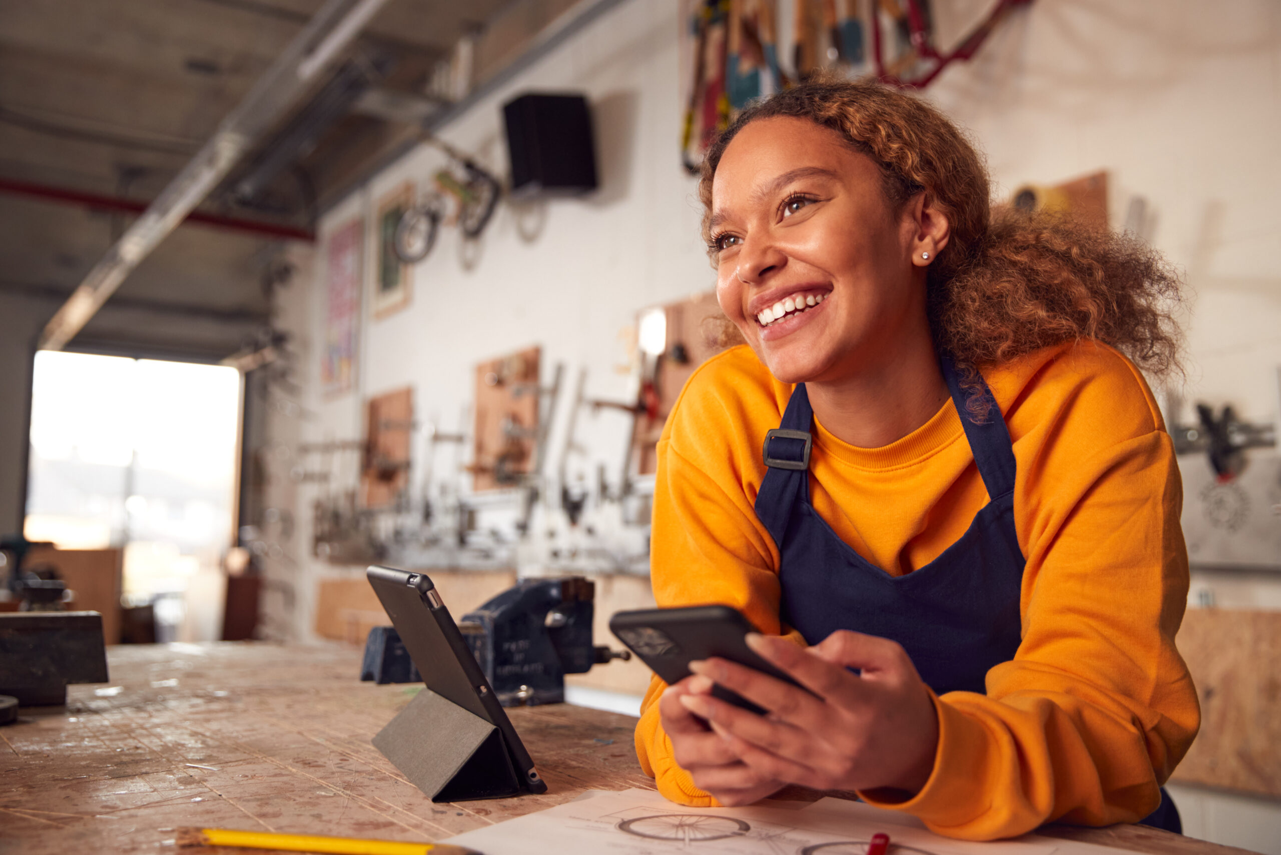 A young woman wearing a yellow sweatshirt and blue apron smiles while holding a smartphone, sitting at a workbench in a workshop. Drawings, a tablet, and tools are visible—she checks her savings on her phone amid creative projects.