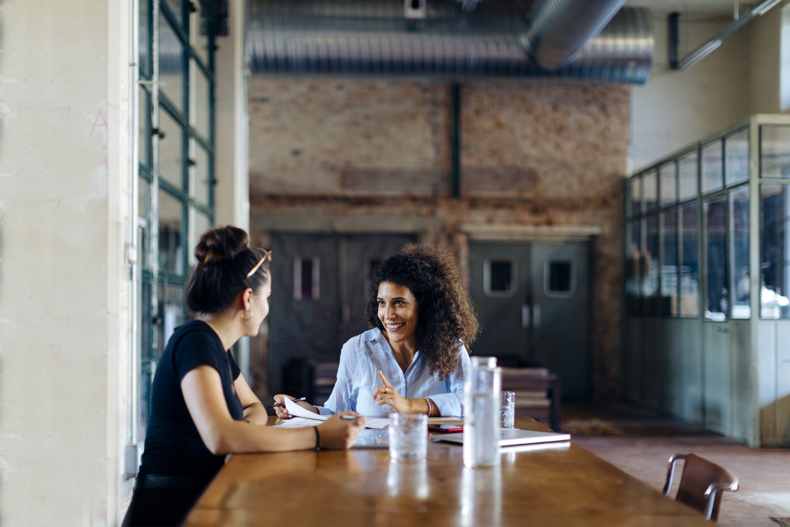 Two women sit across from each other at a wooden table in a modern, industrial-style office, having a friendly like-kind conversation with notebooks and water bottles in front of them.