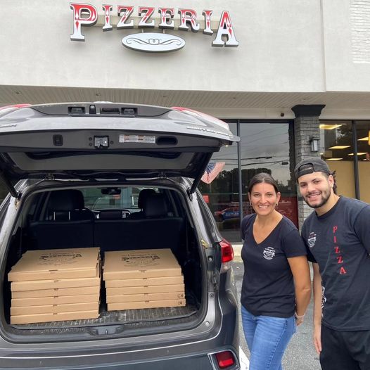 Mariangela and a friend stand and smile beside an open SUV trunk filled with pizza boxes in front of a building with a Pizzeria sign.
