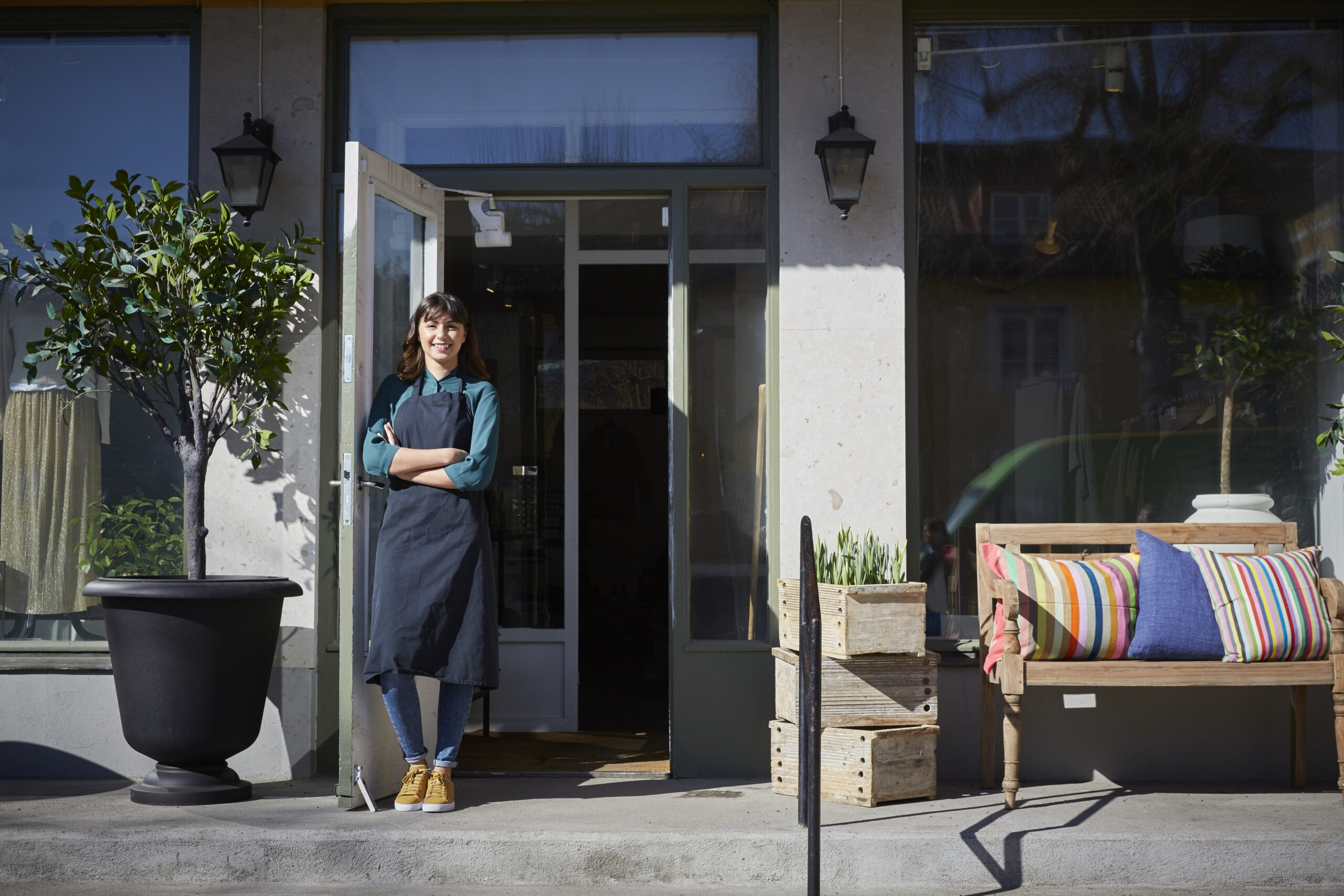 A woman wearing an apron stands smiling with arms crossed in the doorway of her shop, ready to assist with accounting needs. Potted plants and a bench with colorful pillows decorate the entrance on a sunny day.