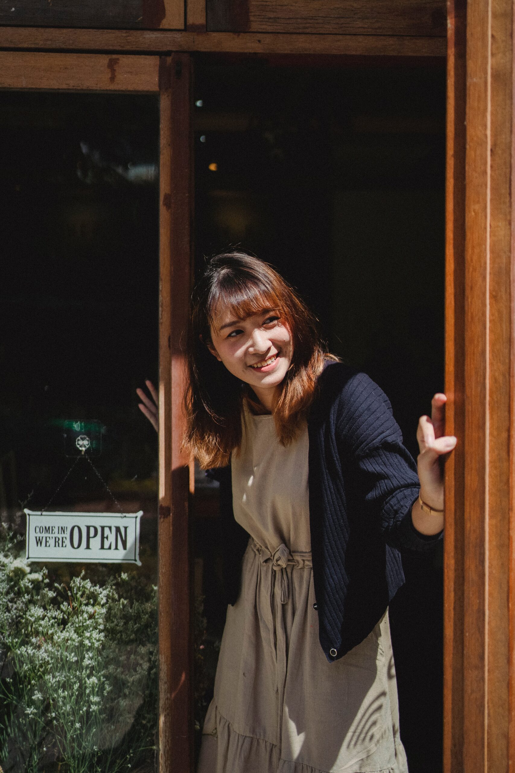 A woman smiling and holding open a wooden door, with a sign that reads Come in, were OPEN visible on the glass. Sunlight shines on her as she stands at the entrance of a shop or cafe.