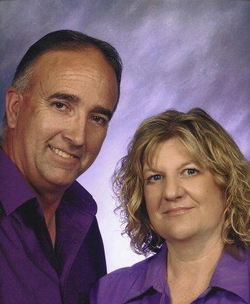 Jerry and a woman, both in purple shirts, pose together in front of a purple and white studio backdrop, smiling softly at the camera.