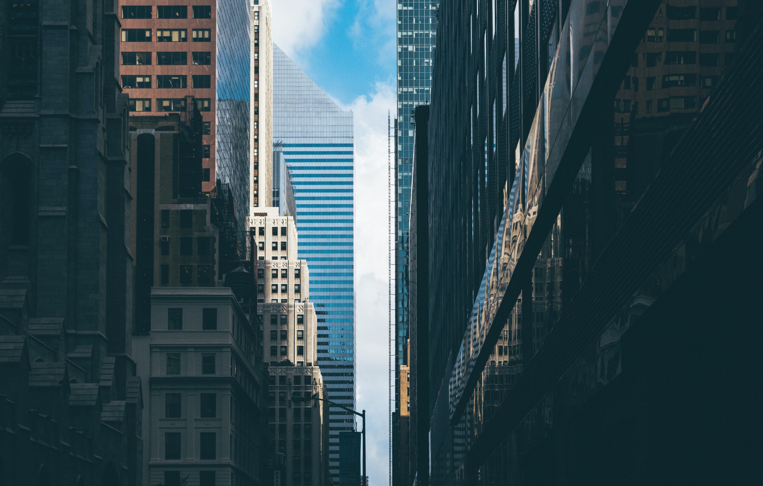 Tall skyscrapers line both sides of a narrow city street, creating a canyon-like effect perfect for quiet deduction. Reflections of buildings shimmer in windows, while a blue sky and drifting clouds appear between the towers in the distance.