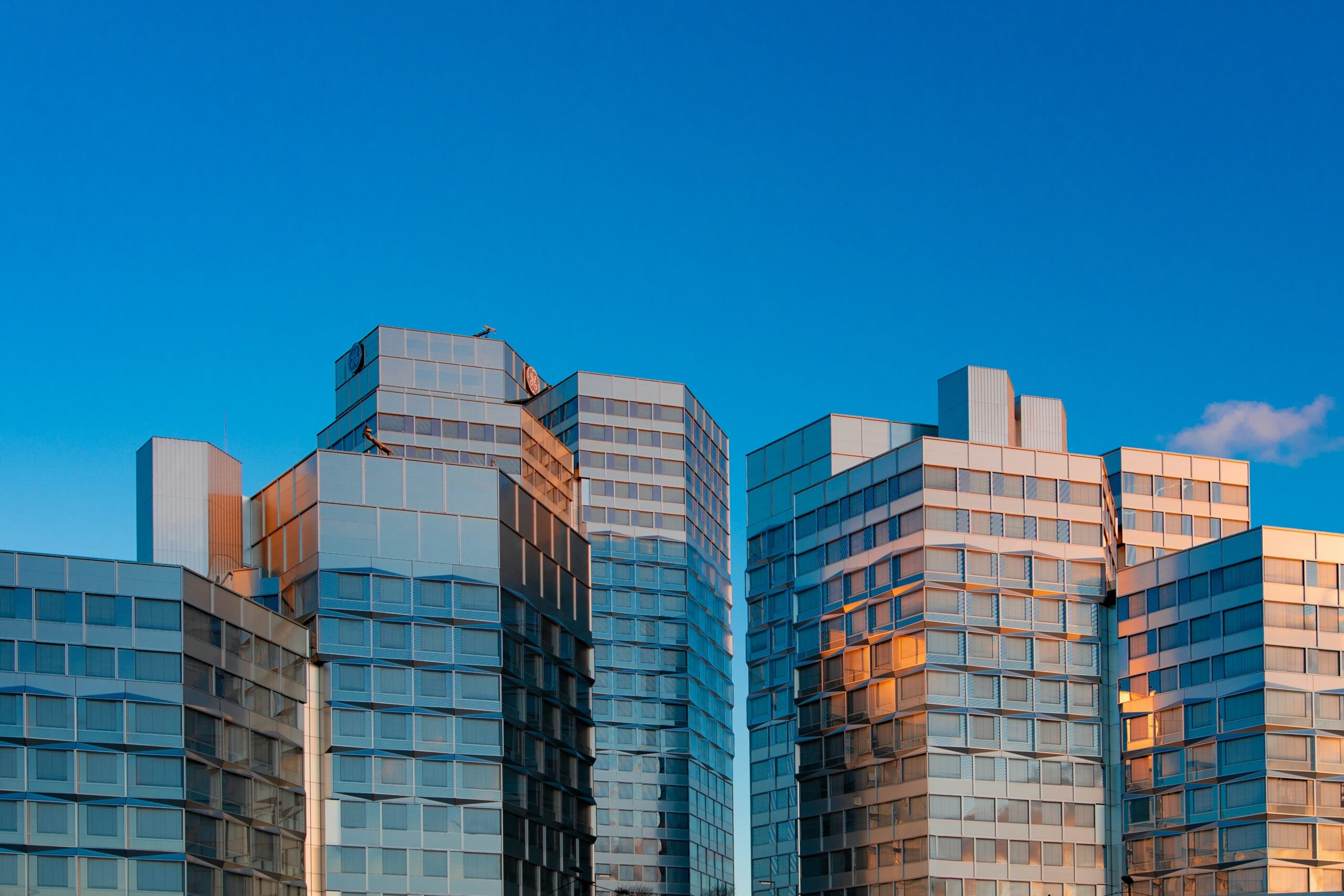 Modern glass office buildings reflecting sunlight, set against a clear blue sky with a few clouds, create an atmosphere where sharp deduction and innovation thrive.