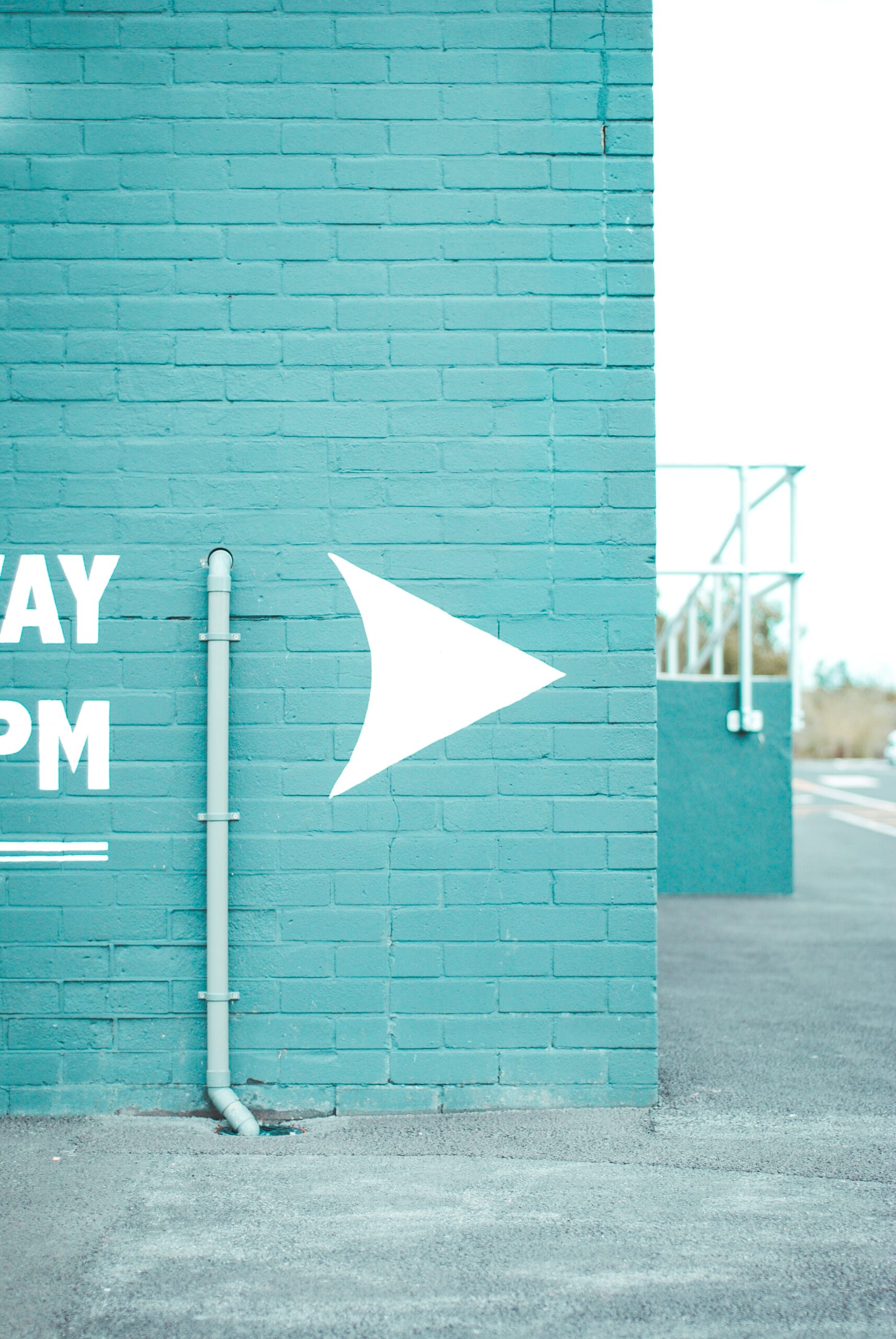 A turquoise brick wall with a white arrow pointing right suggests a pivot ahead. A vertical gray pipe runs up the wall, and partial white text is visible on the left side. The ground is paved, with a railing in the background.
