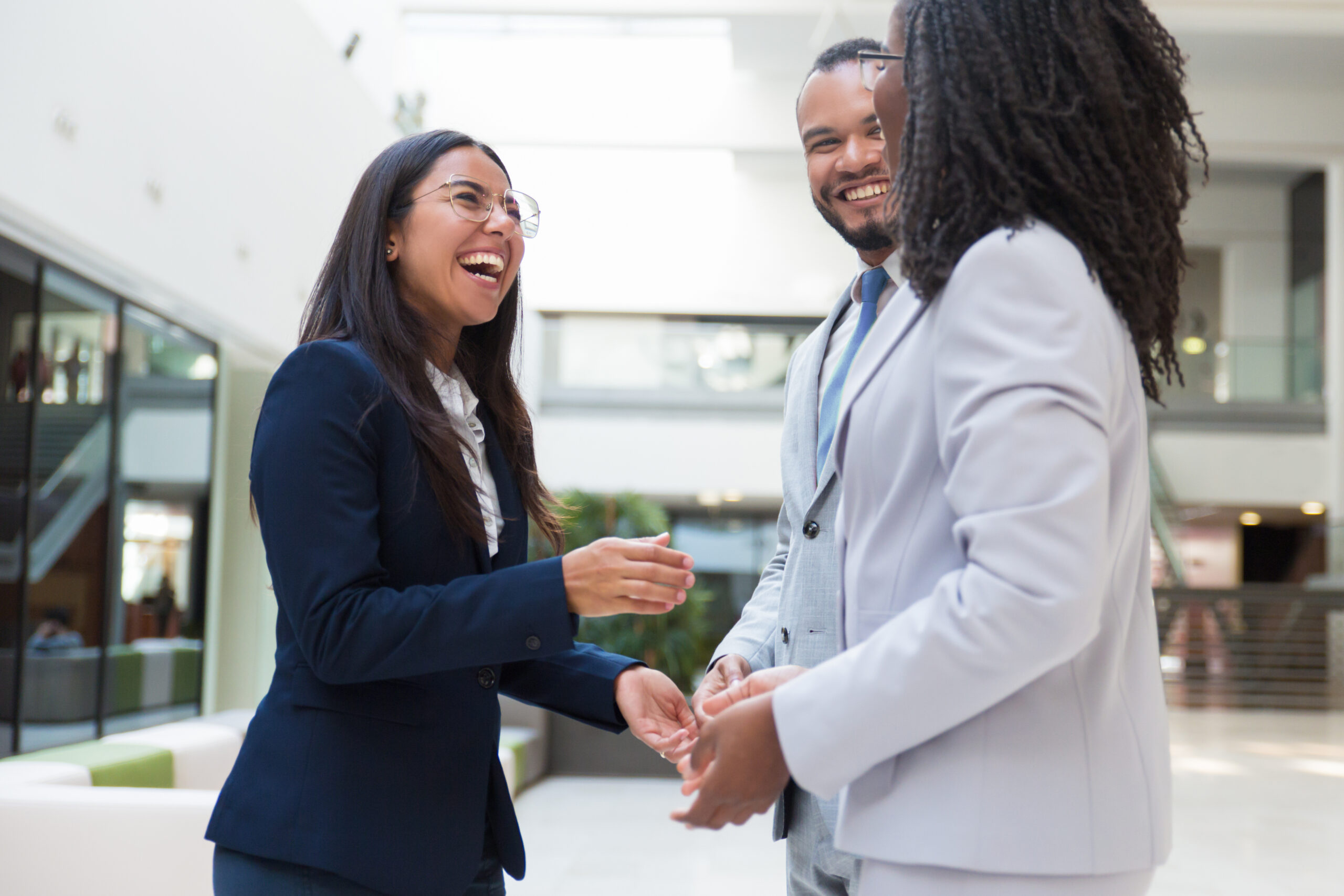 Three professionally dressed people are standing indoors, smiling and talking with each other in a brightly lit office environment.