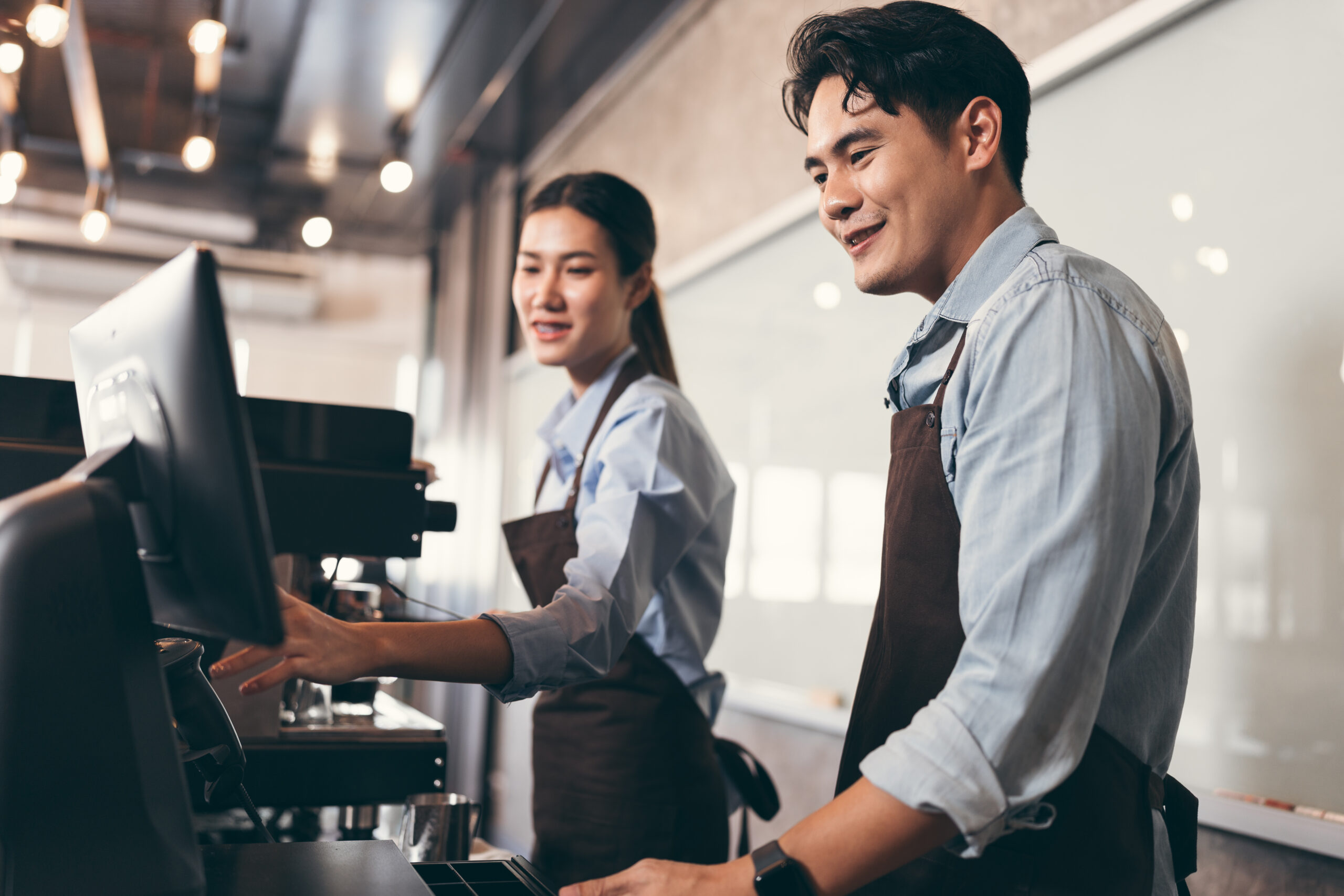 Two partners wearing brown aprons and light blue shirts work at a counter, using a touchscreen register. They are in a modern coffee shop with warm lighting and an industrial interior.