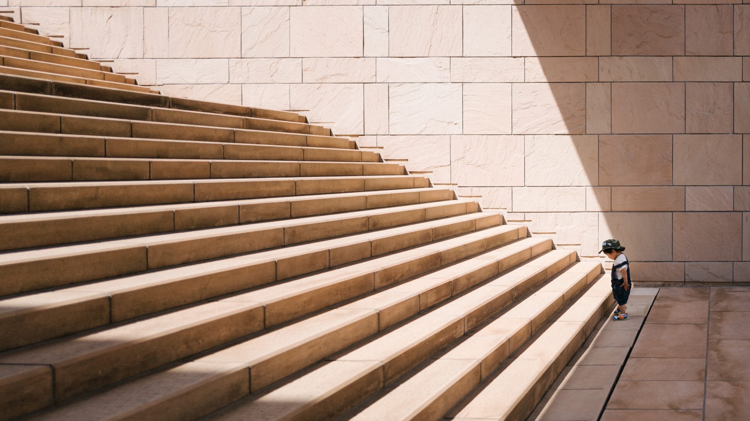 A small child in a hat stands at the base of a large, sunlit stone staircase against a beige tiled wall, gazing up at the towering steps that rise in steady succession.