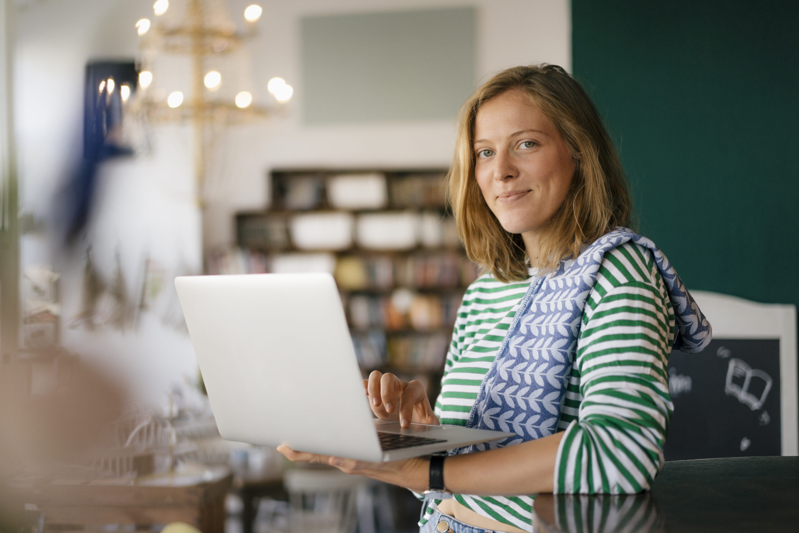 A person in a striped shirt holds a laptop and smiles at the camera in a cozy, well-lit room, perhaps ready to resolve any dispute or challenge that comes their way, with shelves and soft lighting in the background.
