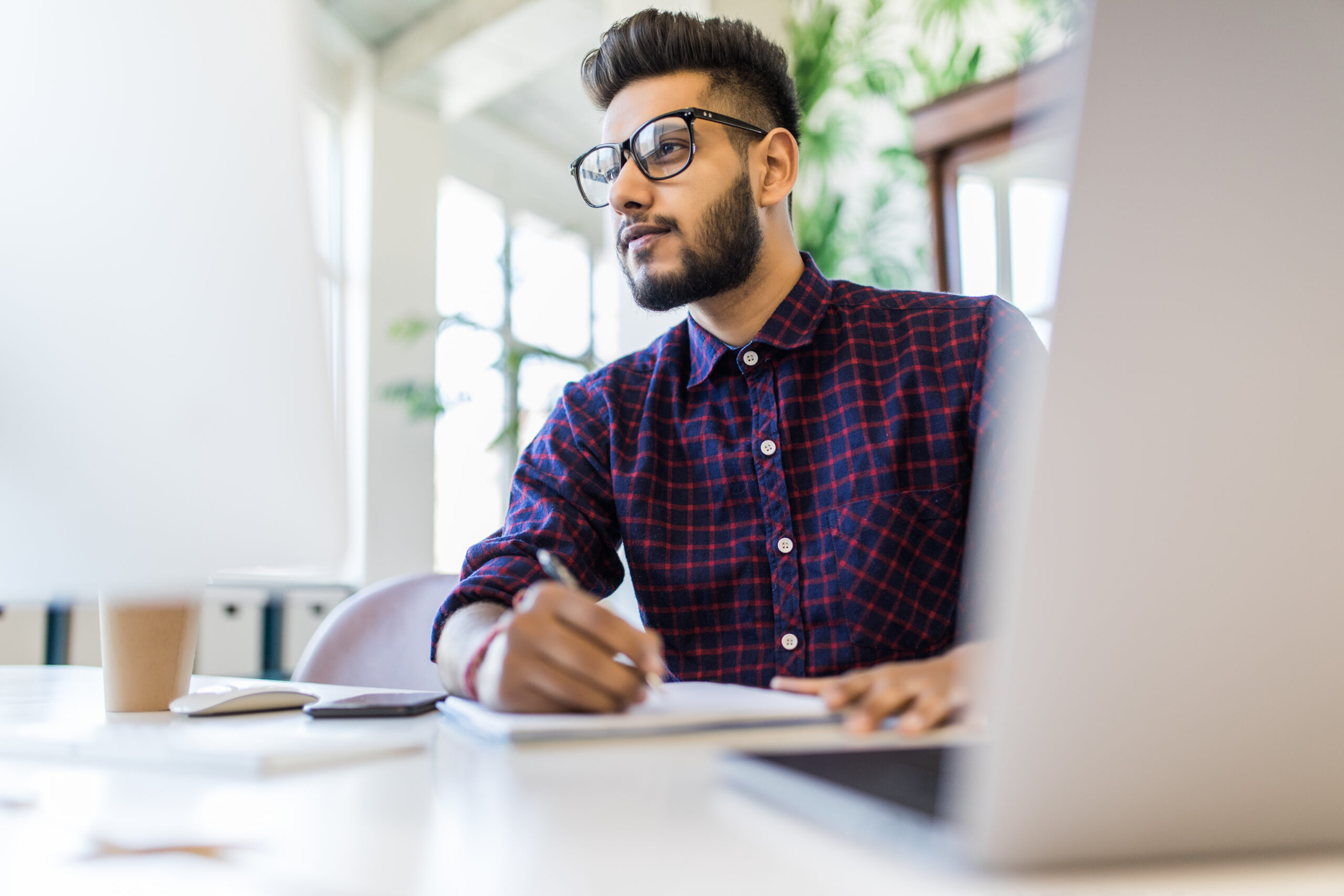 A young man with glasses and a beard, wearing a checkered shirt, sits at a desk writing in a notebook, looking thoughtfully at a computer screen in a bright, modern cyber office space.
