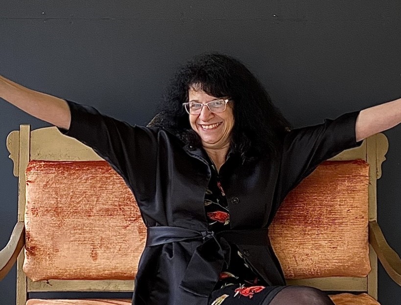 Ann, a woman with curly dark hair and glasses, smiles broadly while sitting on an orange vintage couch, arms outstretched in her black outfit against a dark background.