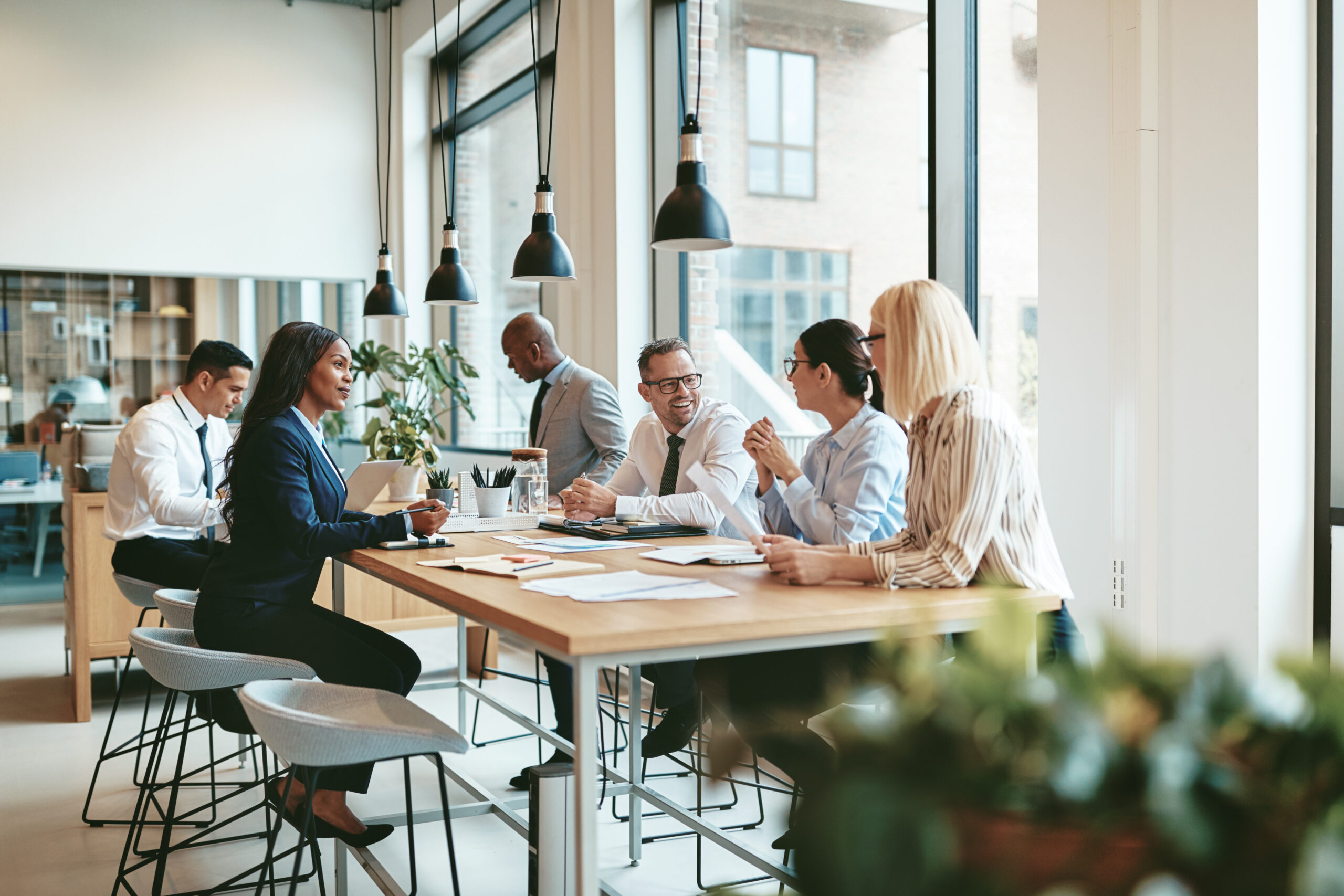 A diverse group of business professionals sits around a large table in a modern, sunlit office, engaged in conversation and collaborating on laptops and documents as they strategize for growth over coffee.