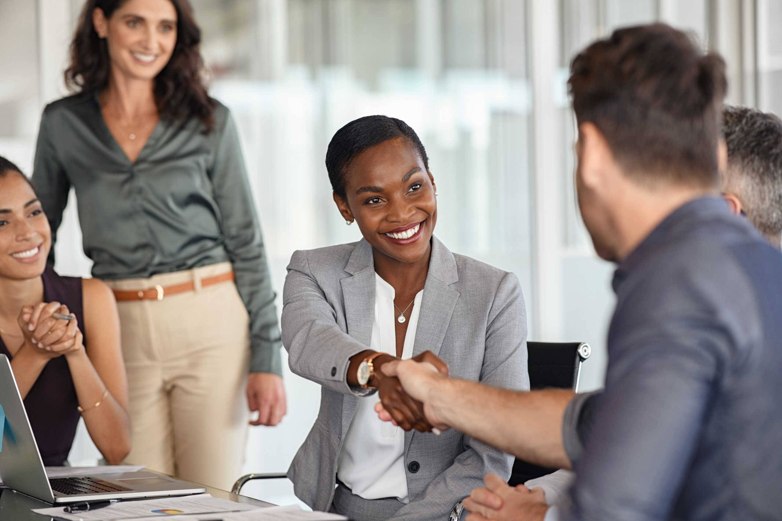 A businesswoman in a gray suit smiles while selling an idea, shaking hands with a man across a conference table as three colleagues look on and smile in a bright, modern office setting.