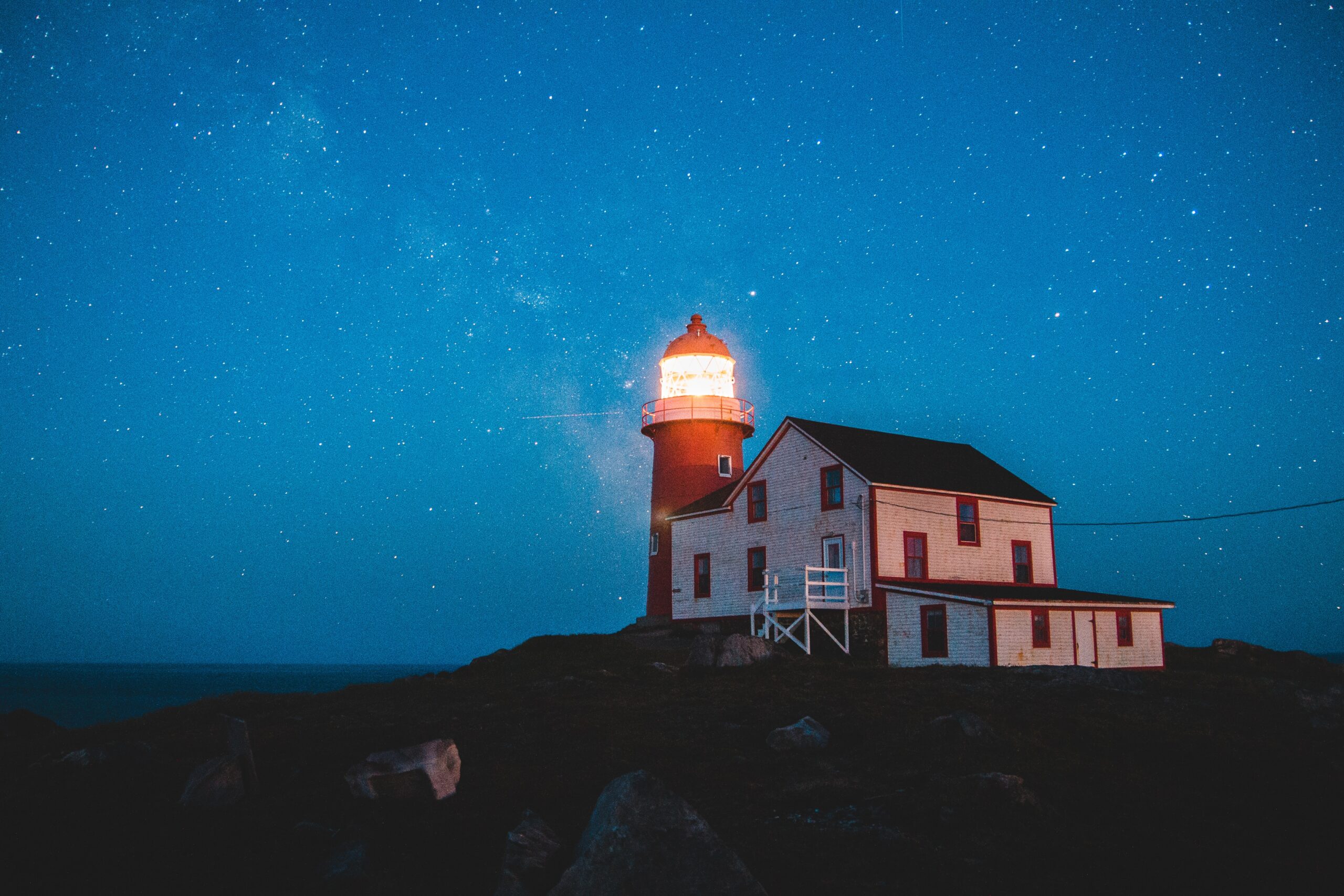A brightly lit lighthouse stands beside a white house on a rocky hill under a starry night sky, its warm glow in the darkness symbolizing innovation guiding the way forward.