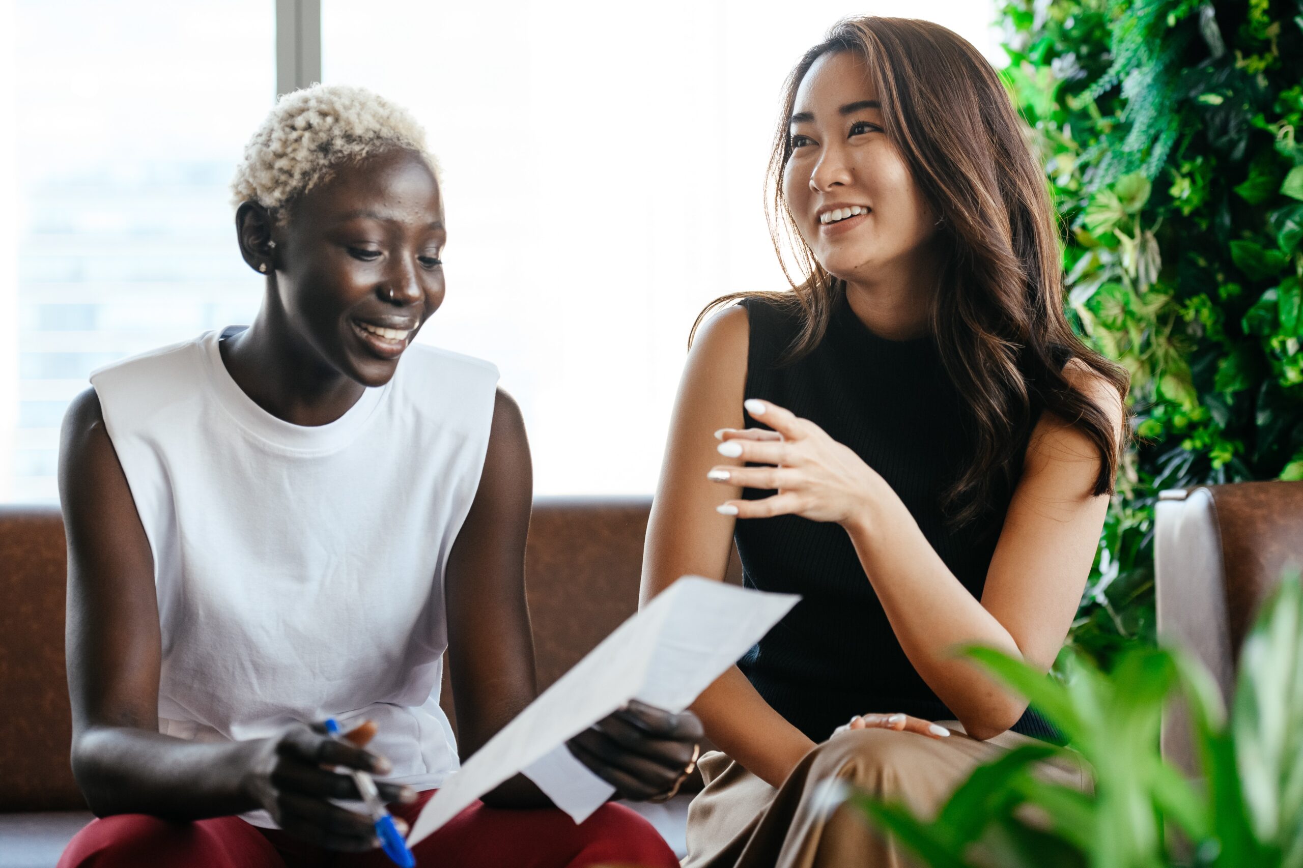 Two women sit together on a couch, smiling and discussing an agreement. One holds a pen and paper as the other gestures, surrounded by green plants in the foreground and background.