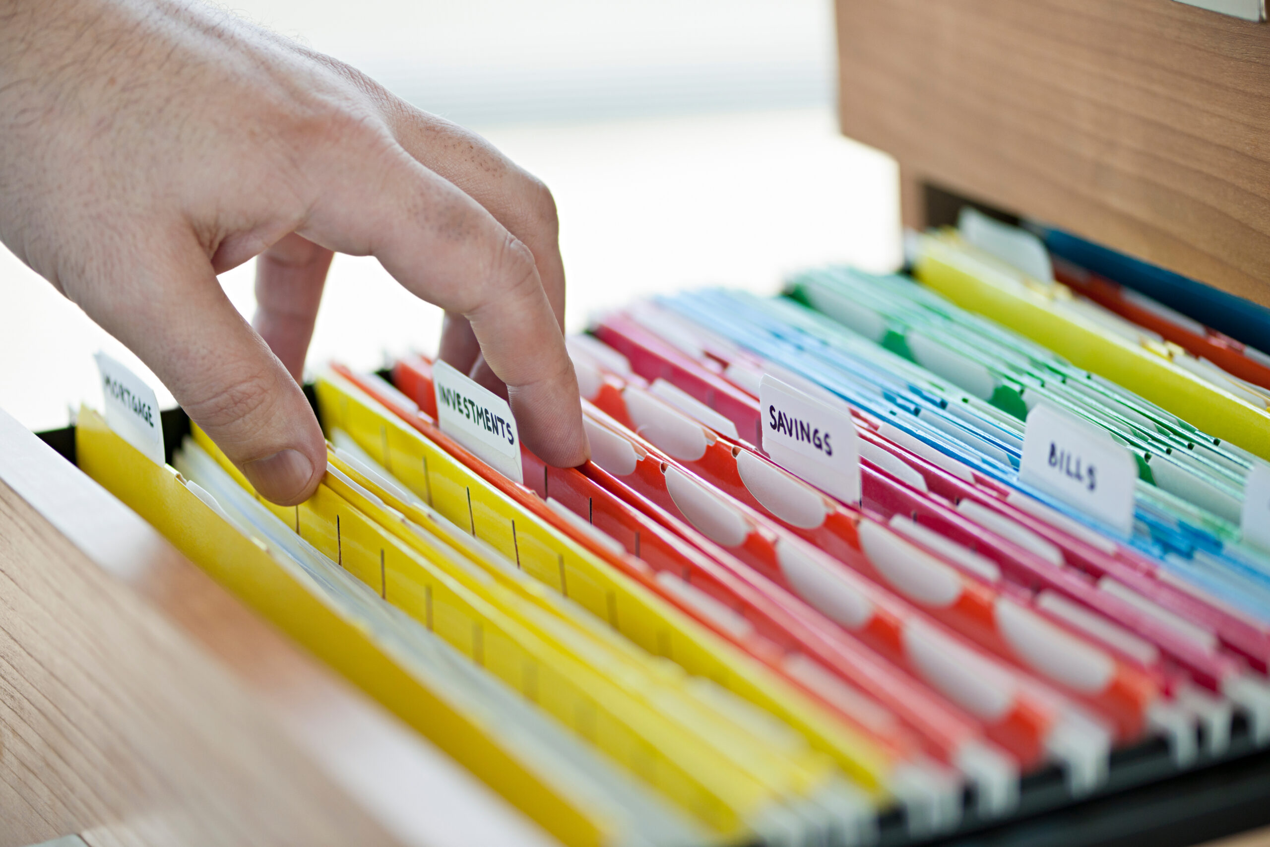 A hand reaches toward color-coded file folders in a drawer, each labeled with categories such as MORTGAGE, INSURANCE, SAVINGS, and BILLS—perfectly organized for efficient year-end review and planning.