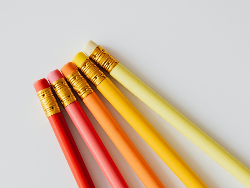 Five business pencils in shades of red, orange, yellow, and cream are arranged in a row on a white surface, with their erasers facing up.