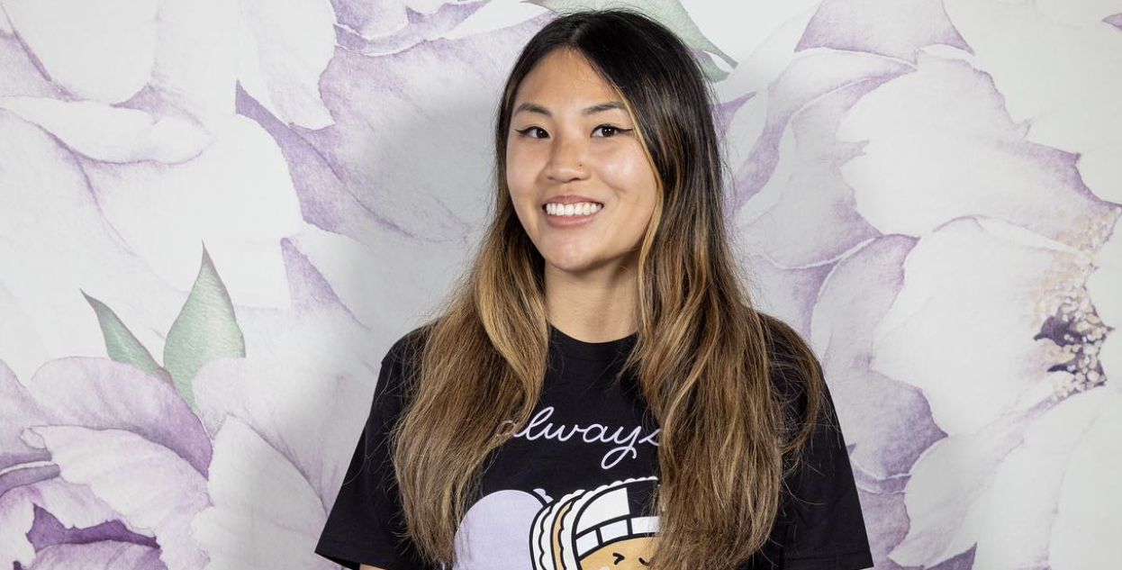 A woman with long brown hair smiles at the camera, standing in front of a wall with a purple and white floral pattern. Radiating success, she wears a black T-shirt featuring a graphic and white text.
