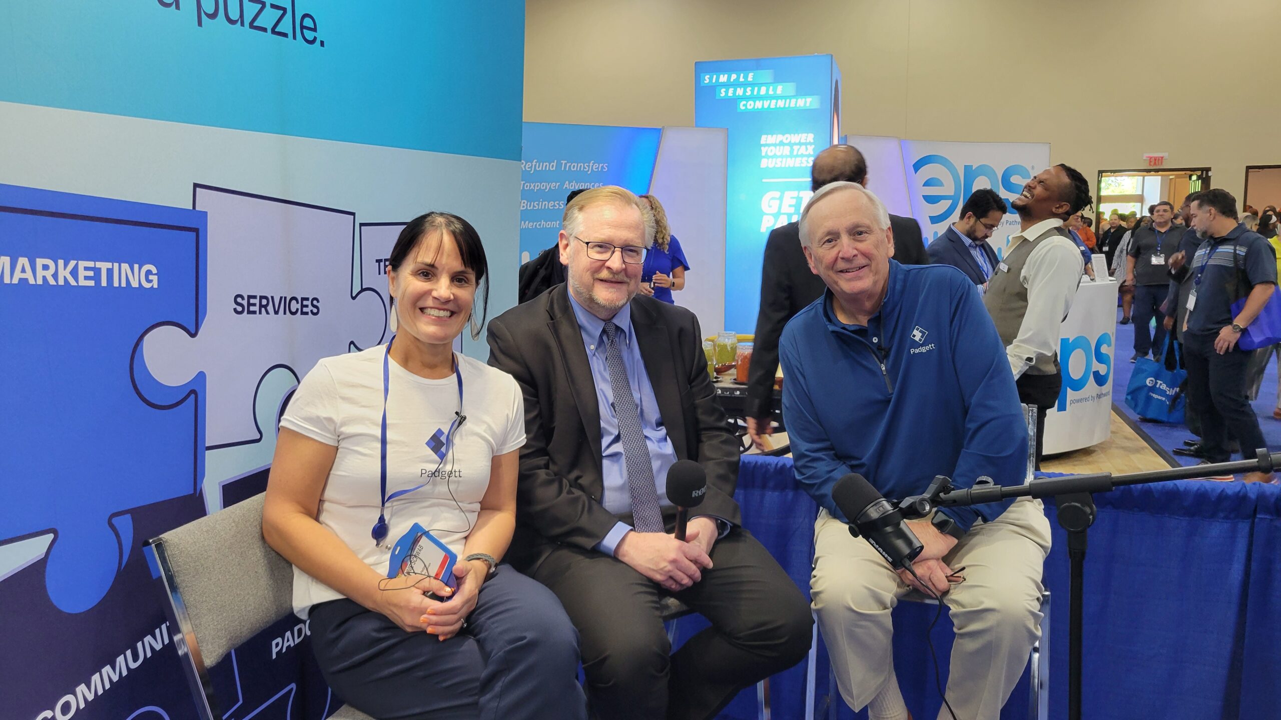 Three people are seated and smiling in front of a blue puzzle-themed backdrop at a conference booth. The woman on the left and the man on the right wear branded shirts, while the man in the center is in a suit and tie.