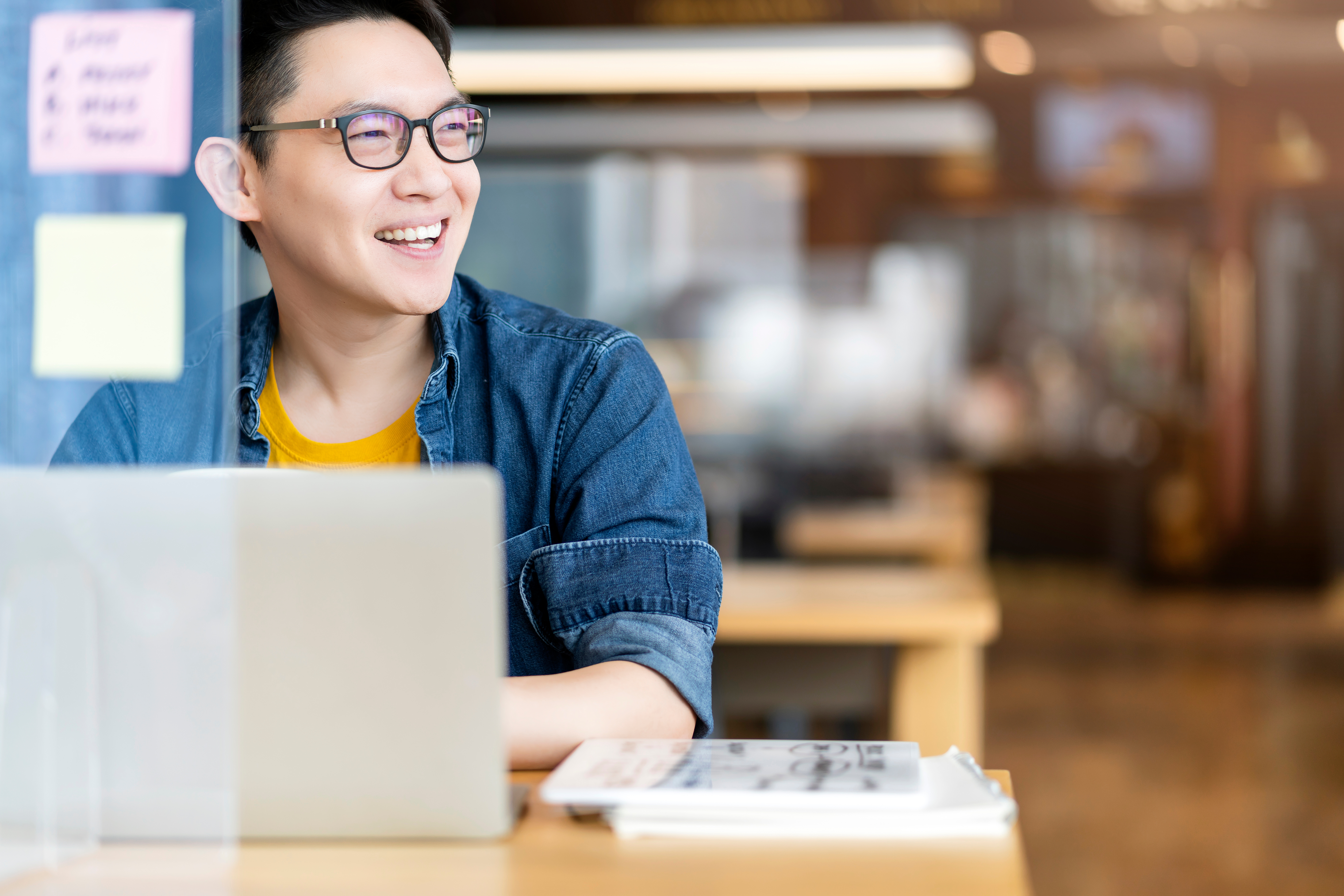 A smiling person wearing glasses and a denim shirt sits at a desk with a laptop and notebook, appearing deep in thought about sales strategies in a bright, modern indoor setting.